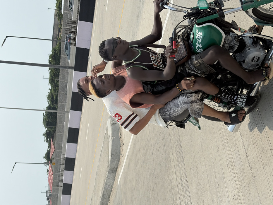 On a clear, sunny day in Jakande, Nigeria, three males are depicted riding a green and black motorcycle on a paved, multi-lane road.
The driver, a dark-skinned male with short dark hair, wears black wraparound sunglasses, a black basketball jersey with "BULLS" text, dark shorts (with "BALENCIAGA" visible), black patterned socks, and black flip-flops. A brown, designer-style crossbody bag is slung over his right shoulder. He grips the handlebars of the motorcycle.
Seated directly behind the driver is the middle passenger, a lighter-skinned male with short bleached blonde hair. He wears a light orange sleeveless top, dark shorts, and a yellow beaded bracelet on his left wrist. He is looking towards the left of the frame, with his right hand raised near his temple.
The rear passenger, a lighter-skinned male with dark dreadlocks, is seated behind the middle passenger. He wears a white short-sleeved t-shirt with "23" on the sleeve, grey patterned shorts, and black slide sandals.
The motorcycle itself is green and black, displaying "K-TOK" on its fuel tank and a visible Bajaj engine.
The environment consists of a wide, light grey asphalt road with clear white lane markings. A concrete road barrier, painted in alternating black and white sections, separates the road from the background. Several tall, dark streetlights with modern fixtures line the road. In the mid-ground, grey block walls, green foliage, and structures with reddish-brown rooftops are visible. Distant grey vehicles are partially discernable on another section of the road in the far background.