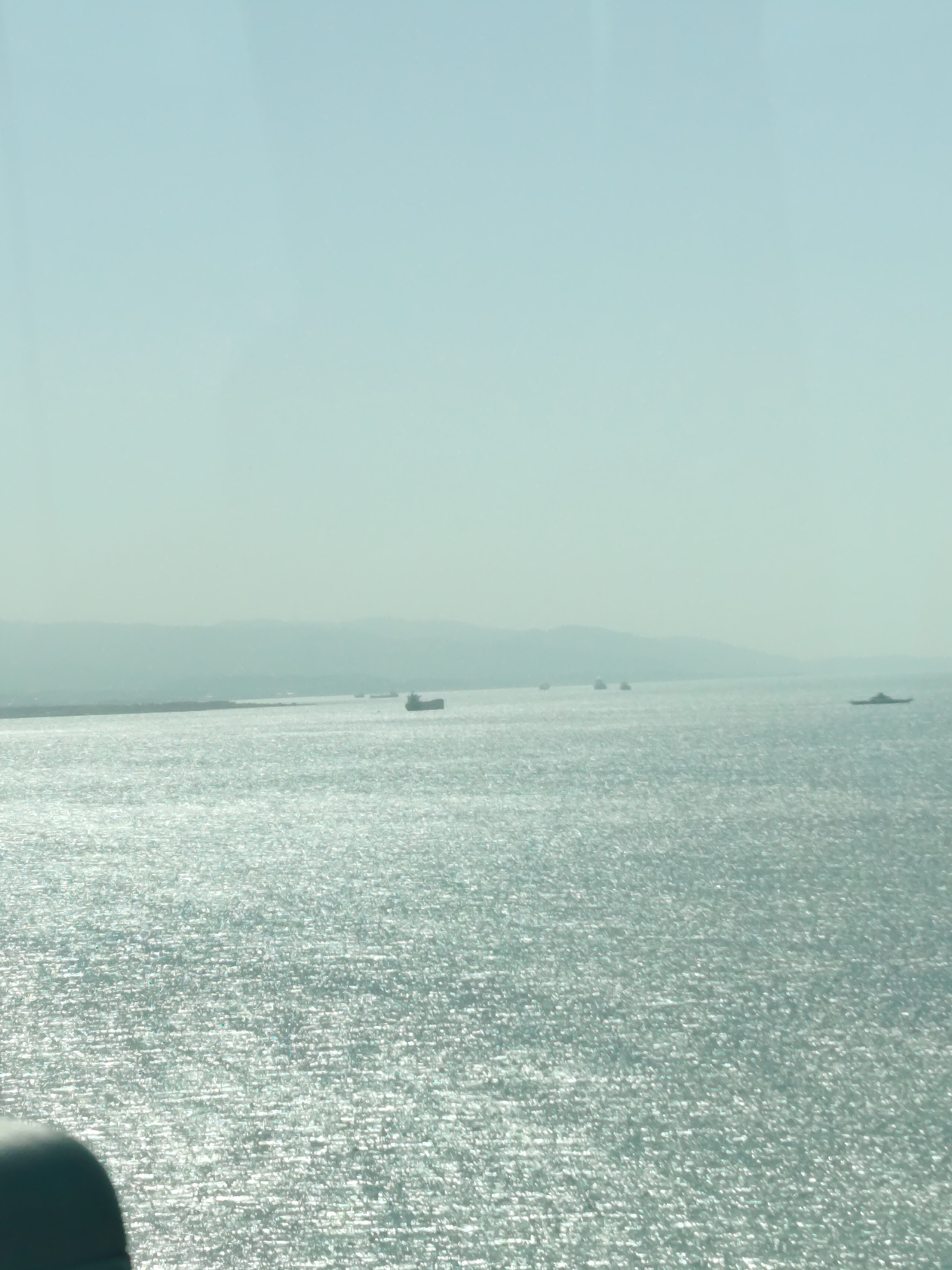 This image captures a wide, expansive view of a large body of water, likely the Gulf of Izmit, consistent with the provided location context of Dilovası, Turkey. The scene is dominated by a bright, shimmering sea under a clear, light blue sky.
In the mid-ground, a hazy distant shoreline with undulating landforms or mountains stretches across the horizon. Several ships are visible on the water. A prominent, dark-hulled vessel, possibly a cargo ship or tanker, is situated slightly right of the center. Further to the left, two smaller, less distinct ships appear in the distance. On the far right, another vessel, appearing flatter and wider, is also present. These ships suggest ongoing maritime activity.
The time of day appears to be bright daylight, possibly midday or early afternoon, as evidenced by the intense sunlight reflecting off the water's surface, creating a sparkling, almost metallic texture across the entire visible expanse. The weather is clear and sunny, though a slight atmospheric haze softens the details of the distant land.
In the bottom-left corner, a dark, indistinct shape partially obscures the water, suggesting the photograph was taken from a structure, vehicle, or a boat's interior. The overall impression is one of calm, vastness, and a clear, bright day on the water. No visible text is present in the image.