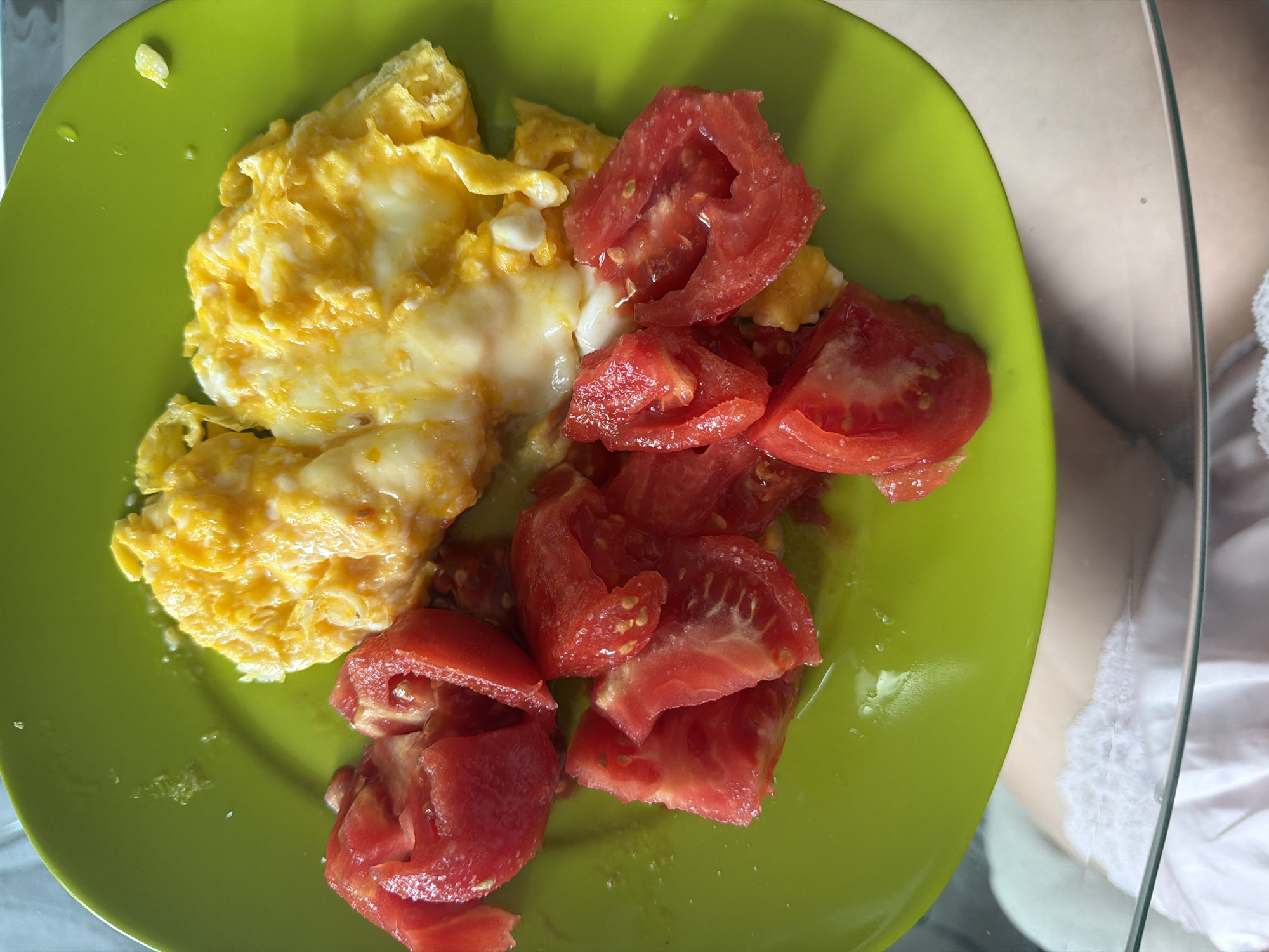 A top-down view shows a bright green plate filled with a meal. The plate contains scrambled eggs and chopped tomatoes. The eggs are a pale yellow with some white curds, appearing soft and fluffy. Scattered among the eggs are several chunks of ripe, red tomatoes, some with visible seeds and juice. The tomatoes appear to be roughly cut, with some pieces larger than others. The plate itself has a slight sheen, and there are a few small droplets of liquid on its surface, particularly around the eggs. The background is slightly out of focus, revealing a light-colored surface, possibly a table, and a hint of what looks like clothing, a white fabric with some texture and a sheer, possibly glass or plastic, surface to the right. The lighting suggests it might be daytime, and the overall scene appears to be a casual meal setting, likely indoors. The location context provided is Sofia, Bulgaria, but there are no specific visual cues in the image to confirm this.