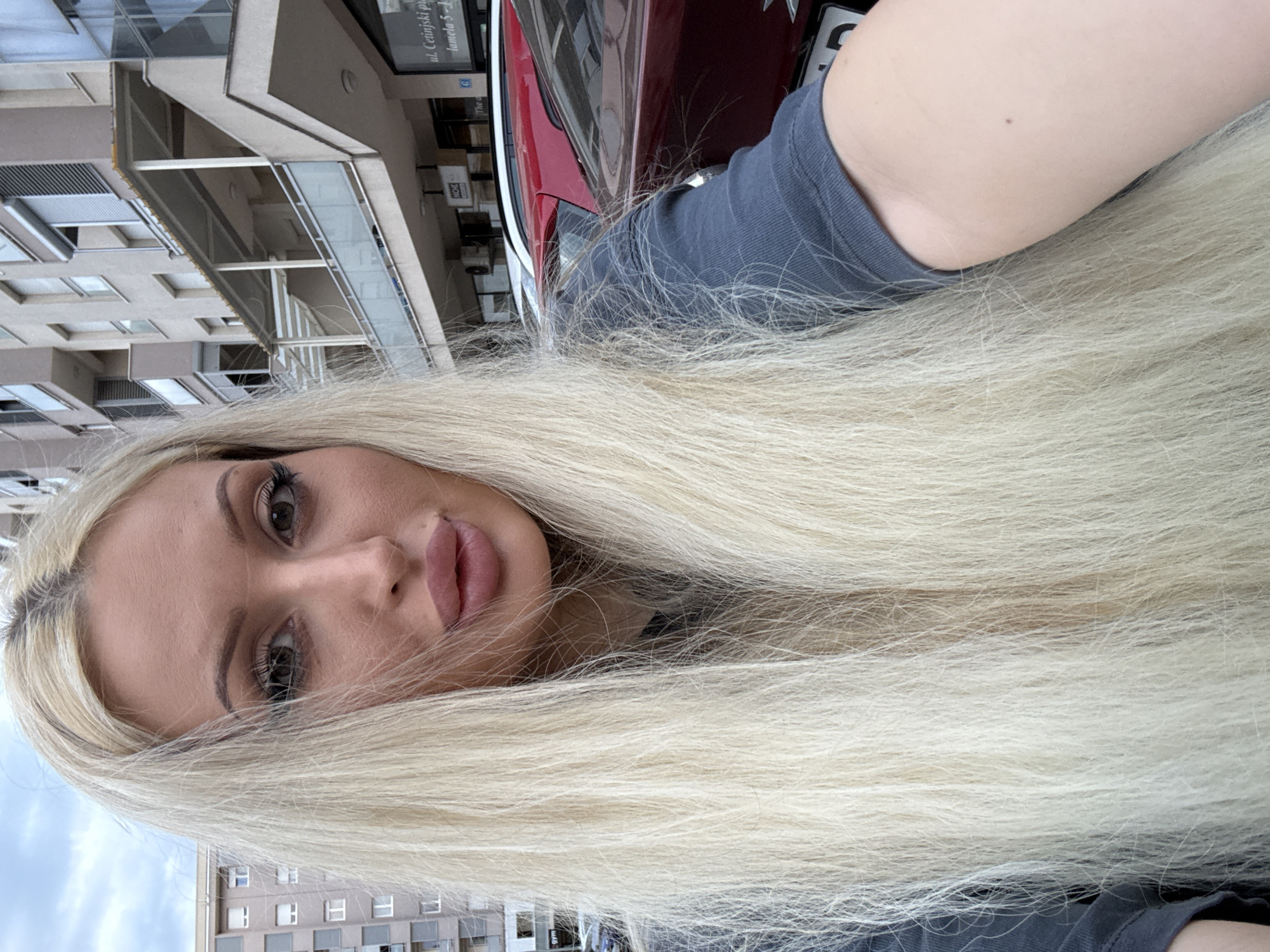 An outdoor, close-up, eye-level selfie shows a blonde woman with long, straight hair. She is wearing a dark gray t-shirt. Her face is tilted slightly upwards, and she is looking towards the camera. Her lips are parted, and she is wearing makeup, including mascara and lip gloss. Behind her, a building with balconies and windows is visible, along with parked cars. The sky is overcast.