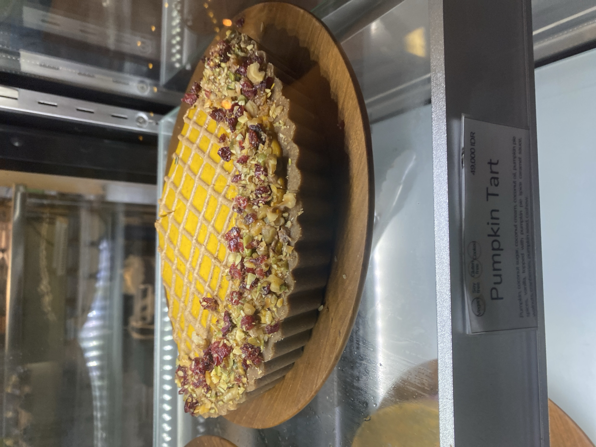A pumpkin tart on a wooden plate is displayed in a glass showcase. A label next to the tart provides details, including the price (49,000 IDR) and a description of the ingredients: pumpkin, coconut sugar, coconut cream, coconut oil, pumpkin pie spices, vanilla, topped with pumpkin pie spice caramel sauce, walnuts, cranberries, pumpkin seeds, and cashews. The label indicates that the tart is vegan, soy-free, and gluten-free. The scene is observed in Ubud District, Indonesia.