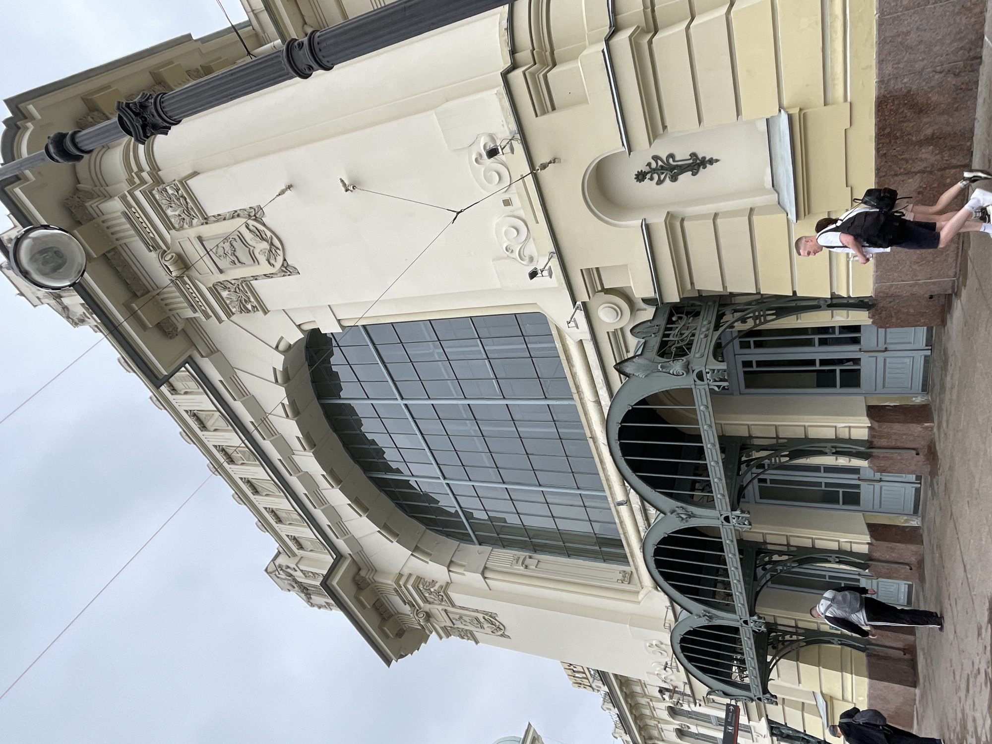 The image depicts the facade of a building in Saint Petersburg, Russia, likely a historic train station due to its grand architecture and the presence of arched entrances. The building is light yellow with ornate white detailing around its large arched windows. A black lamppost with a globe-shaped light fixture stands to the right of the frame. Several people are visible walking on the sidewalk in front of the building. Two individuals are walking away from the camera towards the left, and a man and a woman, both carrying backpacks, are walking towards the right. The sky is overcast.