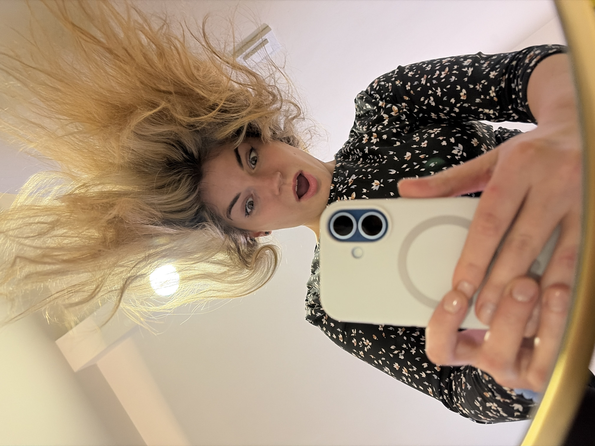 A female individual is depicted in a low-angle mirror selfie, holding a light-colored smartphone towards her reflection. Her long, blonde hair is styled dramatically upwards and outwards, creating a voluminous, spiky appearance. Her facial expression includes wide, open eyes and an open mouth, conveying surprise or a playful shock. She wears a dark, long-sleeved top or dress adorned with a small, light-colored floral pattern. The smartphone, held with both hands, shows its rear panel with a dual-lens camera system, a flash, and a central circular magnetic attachment point on its case. The bottom edge of a golden-yellow mirror frame is visible in the lower foreground. The reflected background consists of a bright, light-colored interior wall and ceiling. Overhead, a circular light source is visible to the right of her head, and a rectangular architectural beam or vent is present in the upper left portion of the reflection. The scene is an interior setting with no specific geographical markers discernible to indicate a location within Budva, Montenegro.