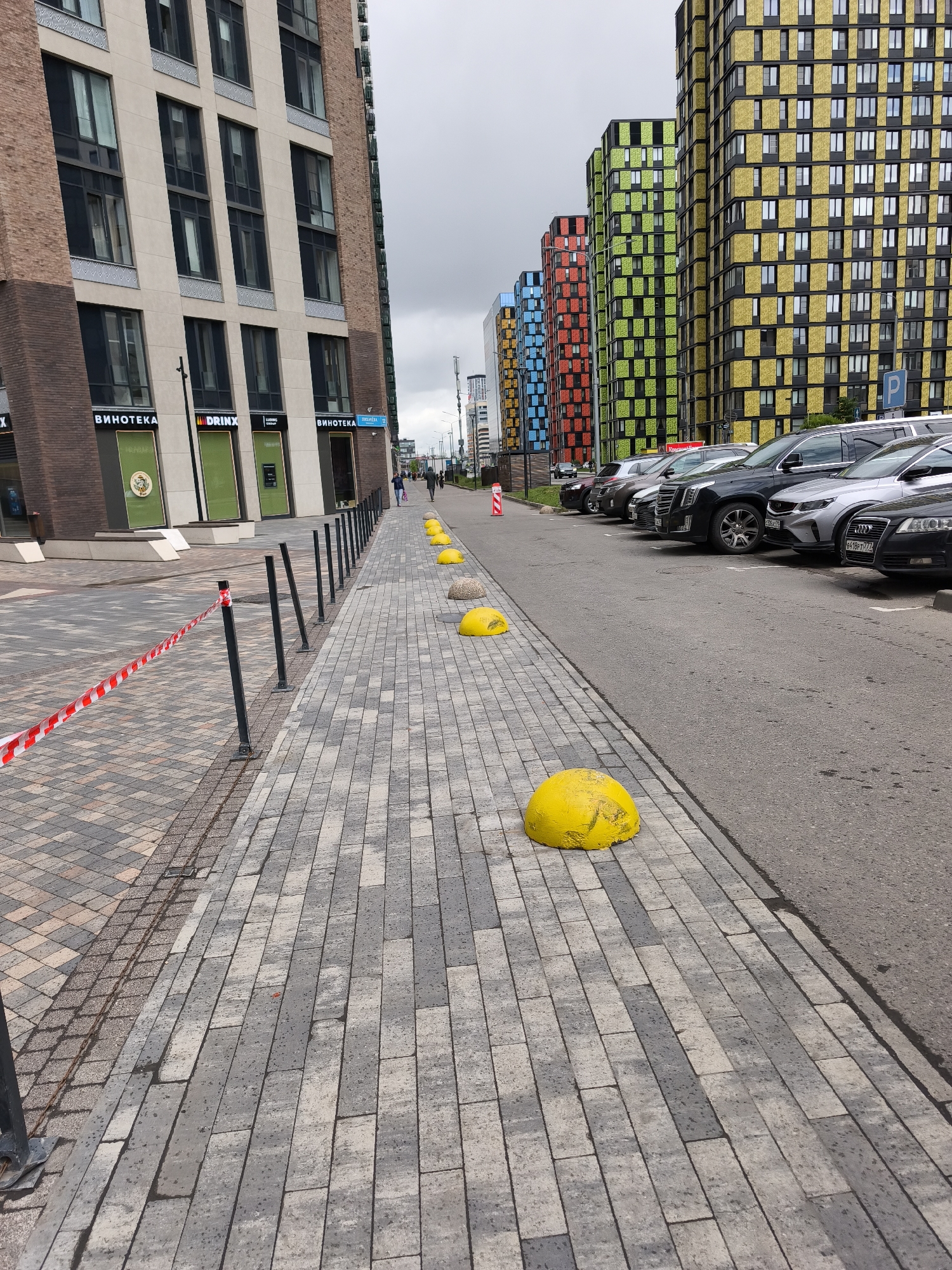 The image depicts a street scene in Moscow, Russia. The perspective is from a pedestrian walkway paved with gray bricks. A line of black metal poles with red and white striped tape runs along the edge of the walkway. Interspersed along the walkway are several yellow, dome-shaped bollards.
To the right of the walkway is an asphalt road with parked cars. The cars are of various makes and models, including an Audi.
Beyond the cars are a series of modern, high-rise buildings. The buildings have distinct, colorful facades, with geometric patterns in shades of blue, red, green, and yellow. The buildings appear to be residential or commercial properties.
Several people are walking in the distance on the opposite side of the road. The sky is overcast. A sign indicates that a "Vinoteka" (wine shop) is located in the lower level of one of the buildings to the left.
