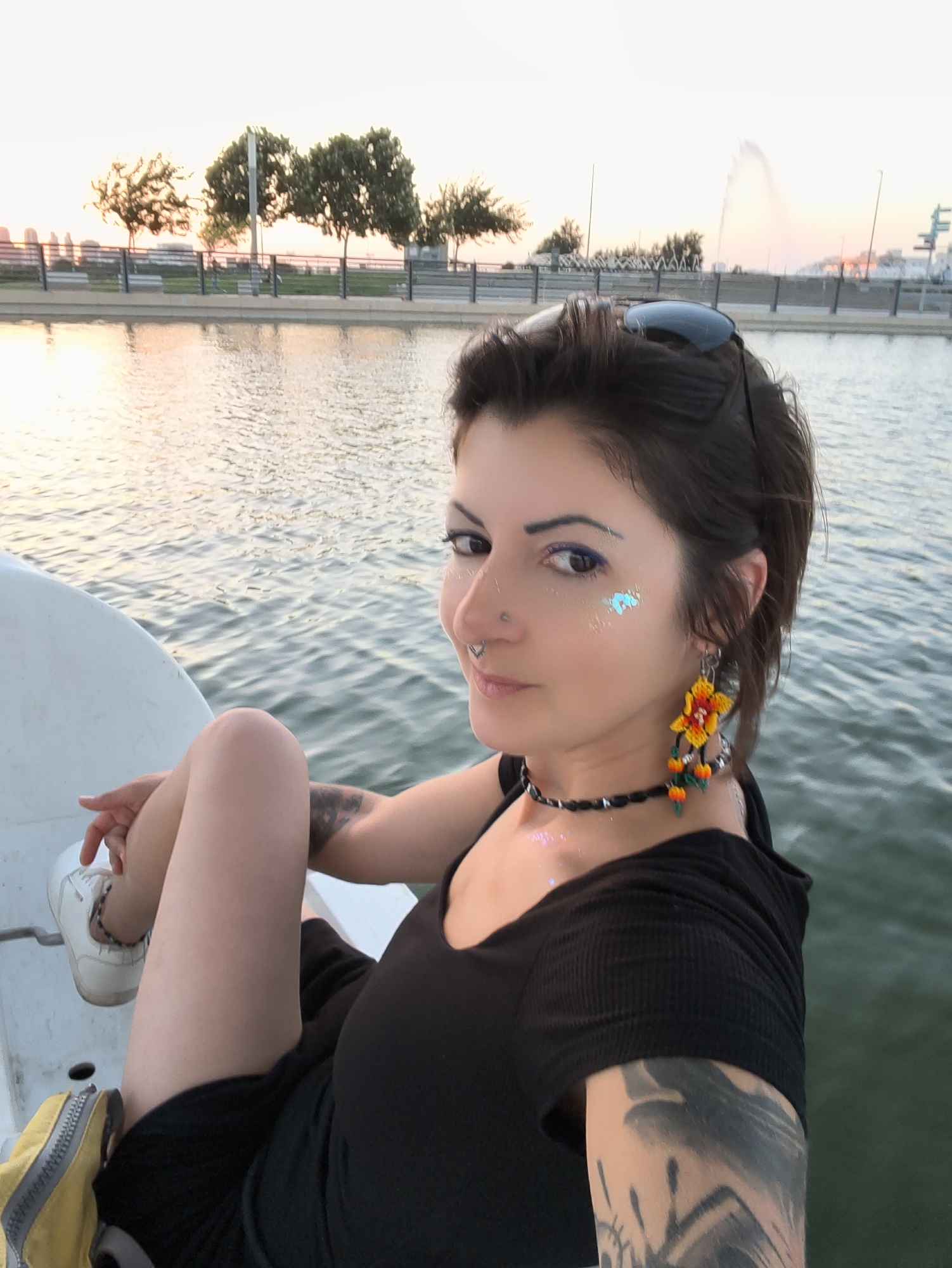 In Be'er Sheva, Israel, a woman with dark hair, styled in loose waves, is pictured sitting on a boat with her left leg bent and resting on her right thigh. She is wearing a black top and black shorts, white sneakers, and a yellow bag is visible near her left leg. Her face is turned towards the camera, with a nose ring, a septum piercing, and glitter makeup on her cheeks and eyelids. She has striking earrings and a choker necklace. The boat is on a body of water, with a city skyline and trees visible in the background. A fountain can be seen in the distance, and the sky is a pale orange, indicating either sunrise or sunset. The water is rippling, reflecting the soft light of the sky. The overall scene suggests a leisurely outdoor activity in a urban park or recreational area.