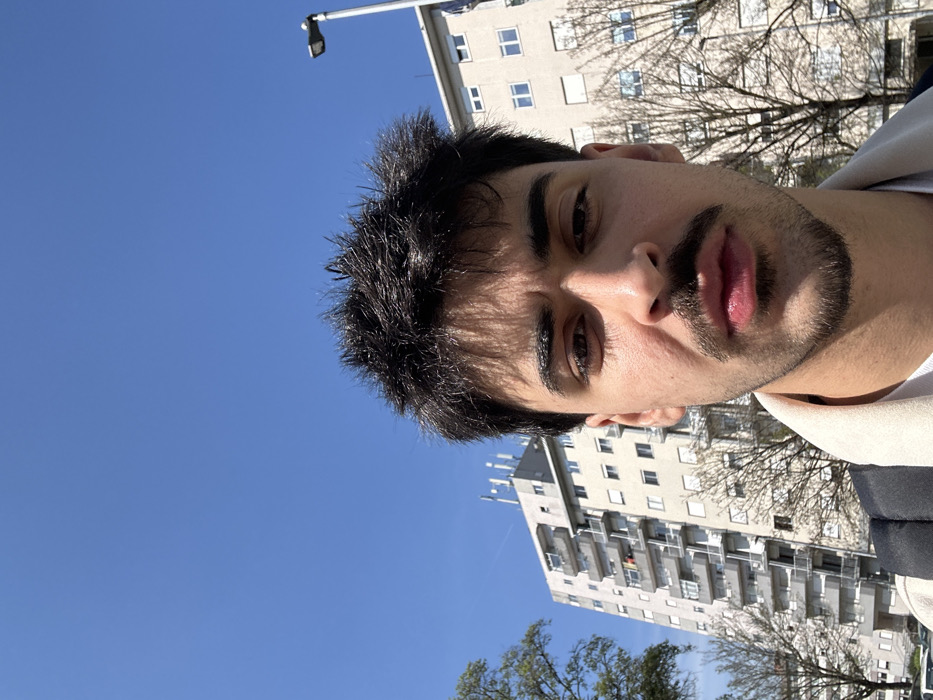 The image depicts a close-up, slightly low-angle portrait of a young adult male outdoors in Guimarães, Portugal, under a clear blue sky. The male subject, centered and facing forward, has dark, spiky hair, dark eyebrows, a mustache, and a short beard. He wears a light-colored collared shirt over a white undershirt, with a dark backpack strap visible over his left shoulder. His expression is neutral.
In the background, to the subject's left, a tall, light-colored multi-story building with numerous balconies and windows is prominent. Clothes are visible drying on at least one balcony, and multiple communication antennas are fixed to the building's roof. To the right of the subject, another similar light-colored building with multiple windows is visible. Several deciduous trees with bare branches are positioned between the foreground and the background buildings. A grey lamppost with a dark fixture extends upwards on the far right side of the frame. The sky is uniformly clear and blue, with a faint white contrail visible in the upper left quadrant. Partially visible at the bottom left are the tops of parked vehicles.