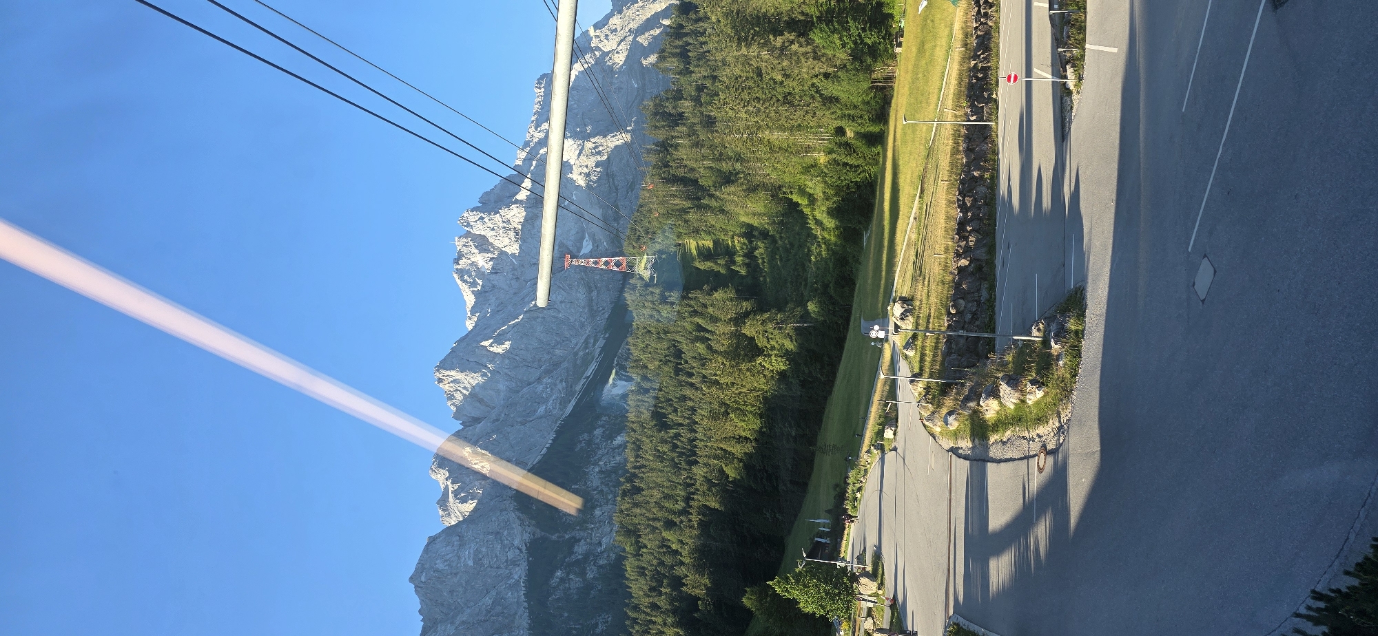 The image shows a mountain landscape with a road in the foreground, in the vicinity of Grainau, Germany. A paved road curves through the scene, with white lines marking parking spaces and lanes. Rocky outcrops and green grass border the road. In the middle ground, a dense forest of green trees covers a hillside. Behind the forest, towering, rugged mountains with snow-capped peaks dominate the background under a clear blue sky. A red and white ski lift tower is visible partway up the mountain slope, with cables extending towards the top. A prominent diagonal light streak, possibly from a window reflection, crosses the upper part of the image. Several street lamps are present along the roadside, and a few people can be seen walking in the distance on the left side of the image. The overall impression is a scenic view of the Alps bathed in sunlight.