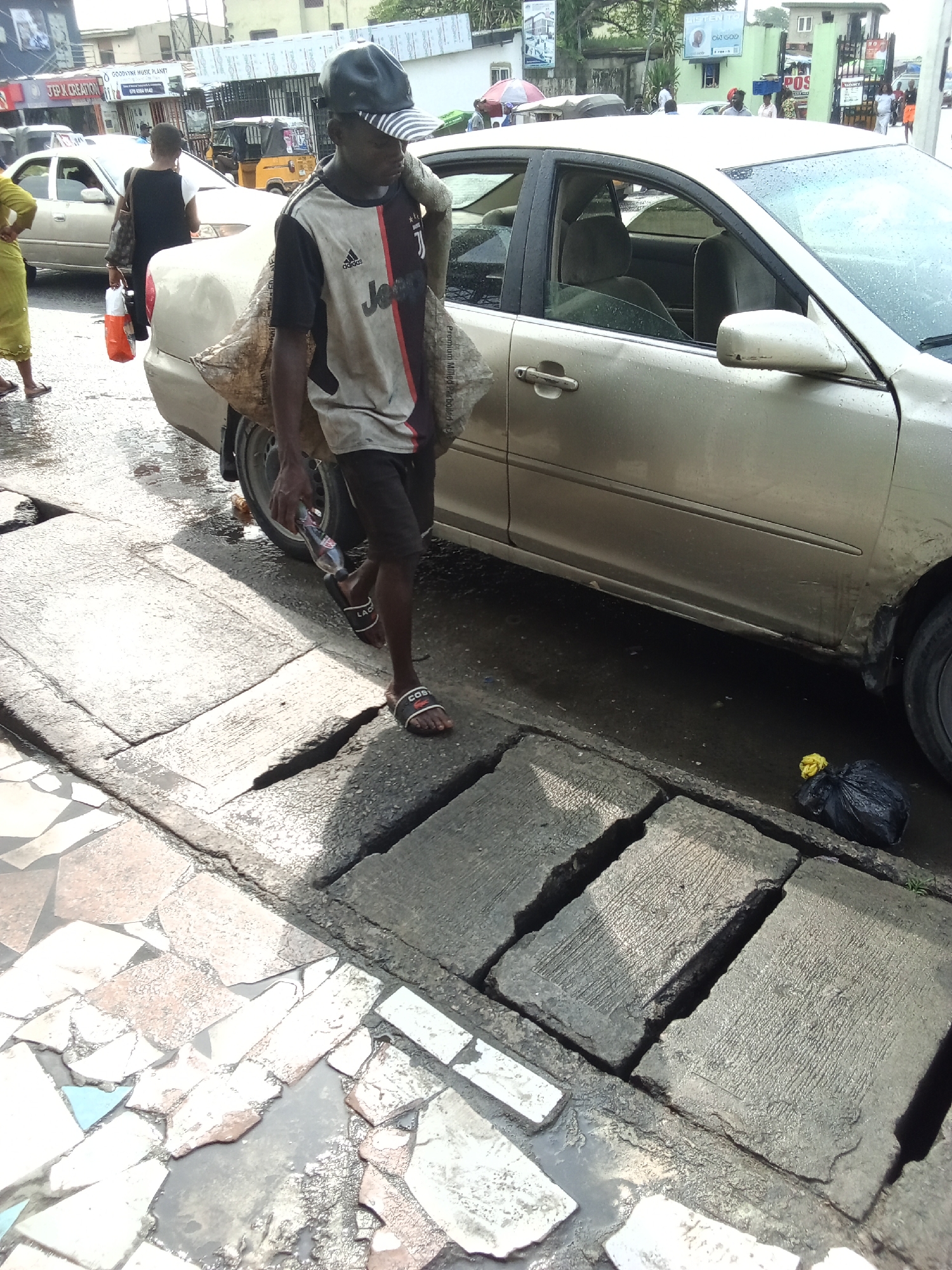 This outdoor street scene, likely captured in Ado, Nigeria, depicts daily life in an urban environment. In the foreground, a man walks on uneven concrete slabs beside a wet road. He wears a striped cap, a two-toned t-shirt with "adidas" and the "JEEP" sponsor logo for Juventus football club, black shorts, and "LAC" (Lacoste) sandals. He carries a large, light-colored sack over his left shoulder and a plastic bottle in his right hand. The ground around him is wet, with puddles reflecting light, suggesting recent rain.
To his right, a gold-colored sedan is parked or moving slowly, its exterior also appearing wet. Further back, another person in a dark outfit with an orange bag is visible near the front of a similar car. On the far left, a person in a light green garment is partially visible on a tiled pavement. In the background, beyond the cars, a yellow Keke Napep (auto-rickshaw) is seen, along with other vehicles and blurred figures of people.
The setting includes various buildings and shop signs. Visible text includes "J.E.P.K CREATION" and "GOODVINE MUSIC PLANET" on shopfronts. A banner or sign in the distance has "OW GOD" (possibly "Our God") and another reads "POS". A small black garbage bag lies by the roadside near the gold car. The overall impression is a bustling street in daylight, with wet conditions after a rainfall.