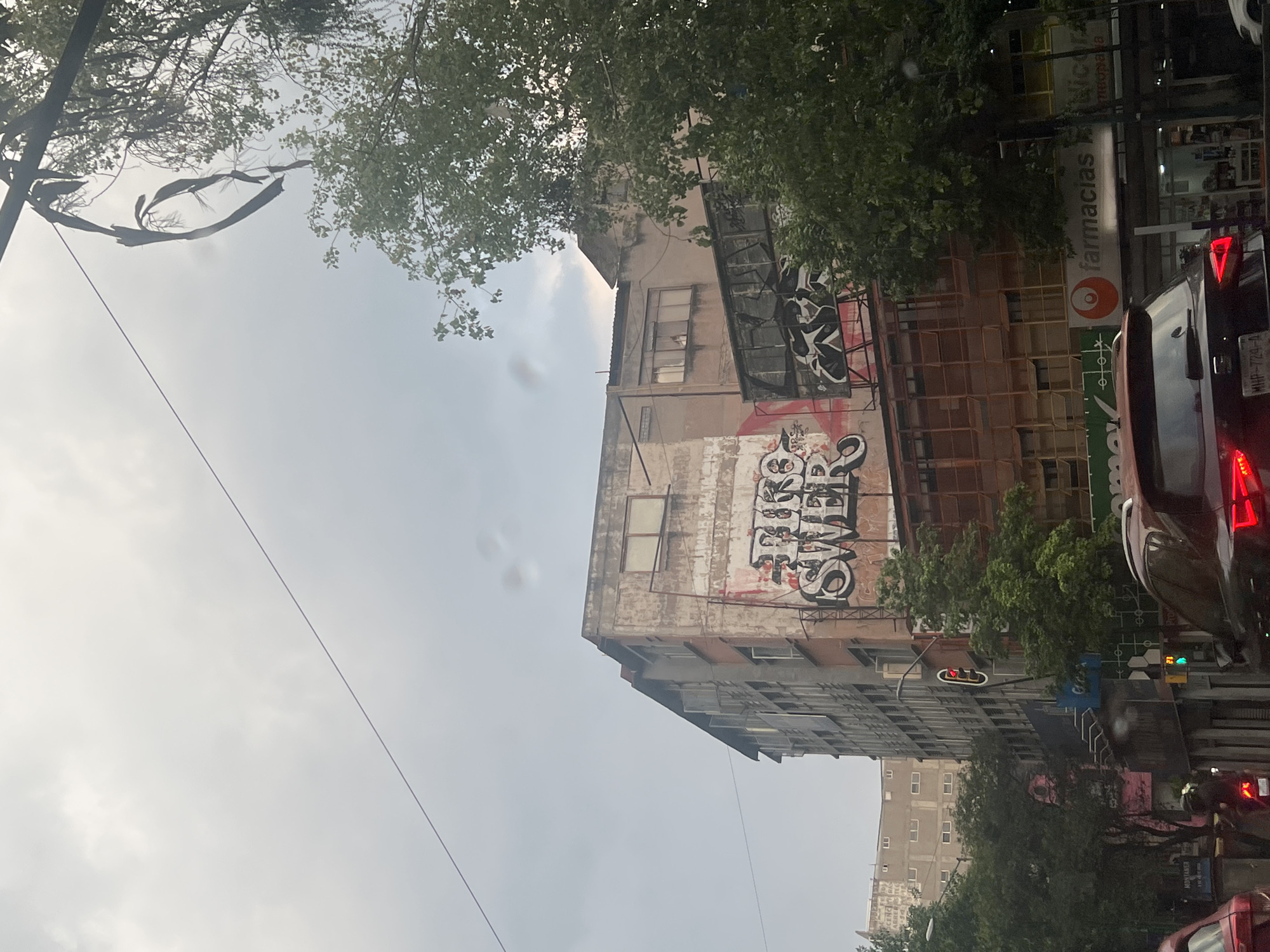 The image captures a street scene in Mexico City, Mexico. A multi-story building with visible graffiti on its exterior is a prominent feature. The building appears to be in a state of disrepair. A “Farmacias” sign is visible adjacent to the building. Street-level activity includes vehicular traffic, with a dark-colored car displaying a license plate number MHP-724-A. Pedestrians are not clearly visible. Overhanging trees partially obscure the view of the building. Overhead cables extend across the frame. The sky is overcast. A traffic light is visible indicating red.