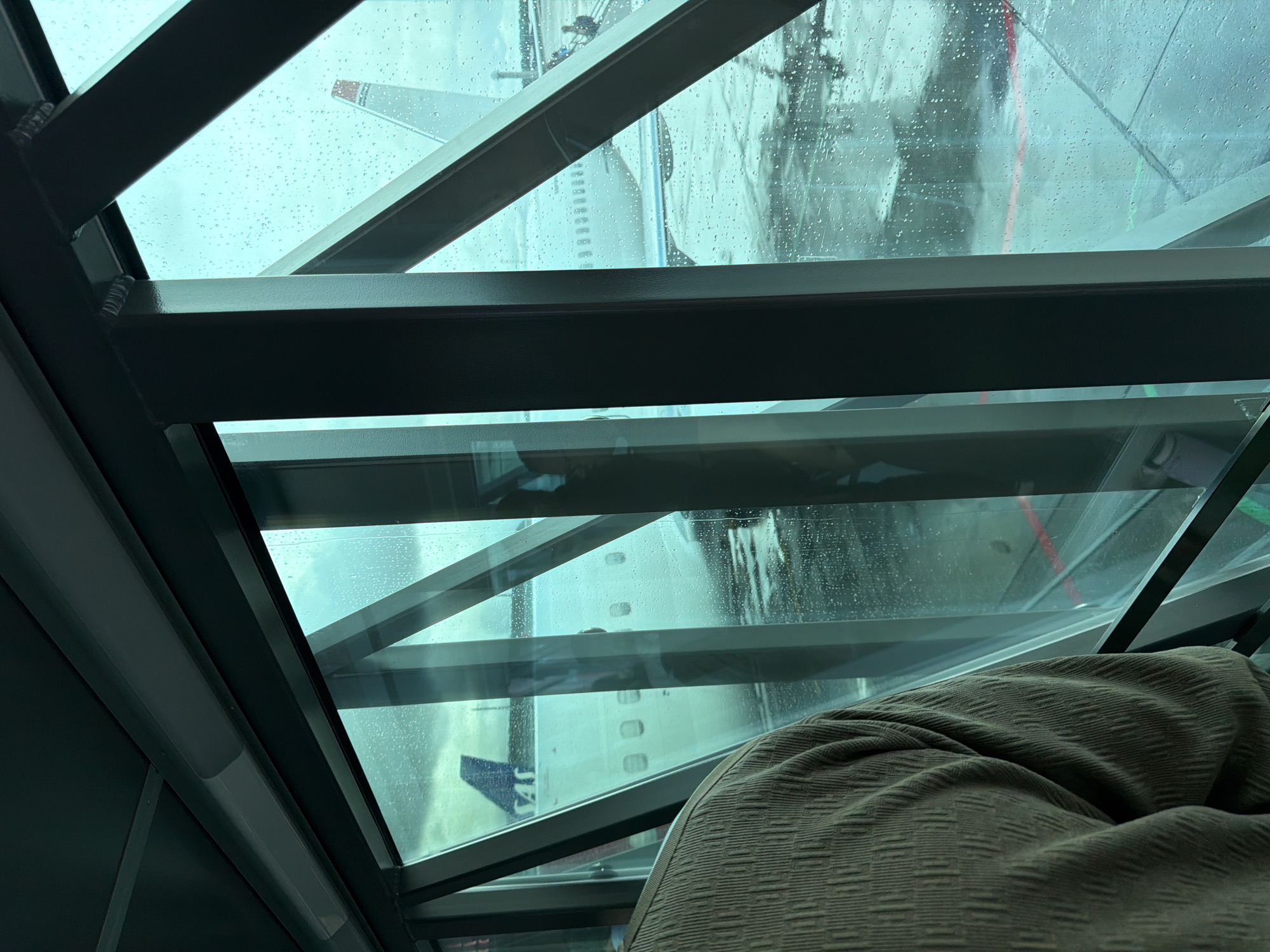 An overhead view looks through a glass jet bridge at an airplane on a rainy day at Gardermoen Airport in Oslo, Norway. The jet bridge has a dark ceiling with a fluorescent light fixture and dark metal support beams. Through the glass, the airplane's fuselage and tail fin are visible, with a SAS logo on the tail. The ground outside is wet and appears to be tarmac, with red and green lines visible. Small water droplets are scattered across the glass. A person wearing a brown, textured sweater is visible in the foreground, on the left side of the image.