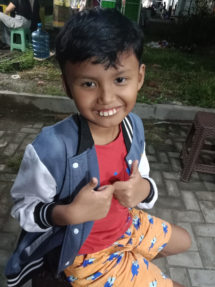 A young male child, estimated to be between 5 and 8 years old, is depicted in a medium shot, centrally positioned, and looking directly at the camera. He has short, dark hair, dark eyes, and tan skin, presenting a wide smile that reveals his upper front teeth. He is wearing a dark blue and white two-tone jacket with white snap buttons over a red t-shirt that has a small blue design on the left chest. Below the waist, he wears orange shorts with blue patterns. Both hands are raised in front of his torso, each forming a thumbs-up gesture. He appears to be kneeling or squatting on a paved surface.

The immediate foreground consists of grey rectangular paving bricks. To the child's left, a person's lower legs, clad in light-colored pants and dark shoes, are visible sitting on a light green plastic stool. Behind this person, a large, translucent blue plastic water bottle rests on the ground. Further left, a yellow sign with partially visible Indonesian text ("CILOR") is mounted on a green structure, likely a food stall. The ground beyond the paved area shows patches of grass and some discarded items. To the child's right, a dark brown, woven plastic stool is present on the paved surface. The background is dimly lit, indicating an evening or nighttime setting, with additional individuals and structures, potentially market stalls, visible in the distance. The scene is located in the city of Turunsih, Indonesia.