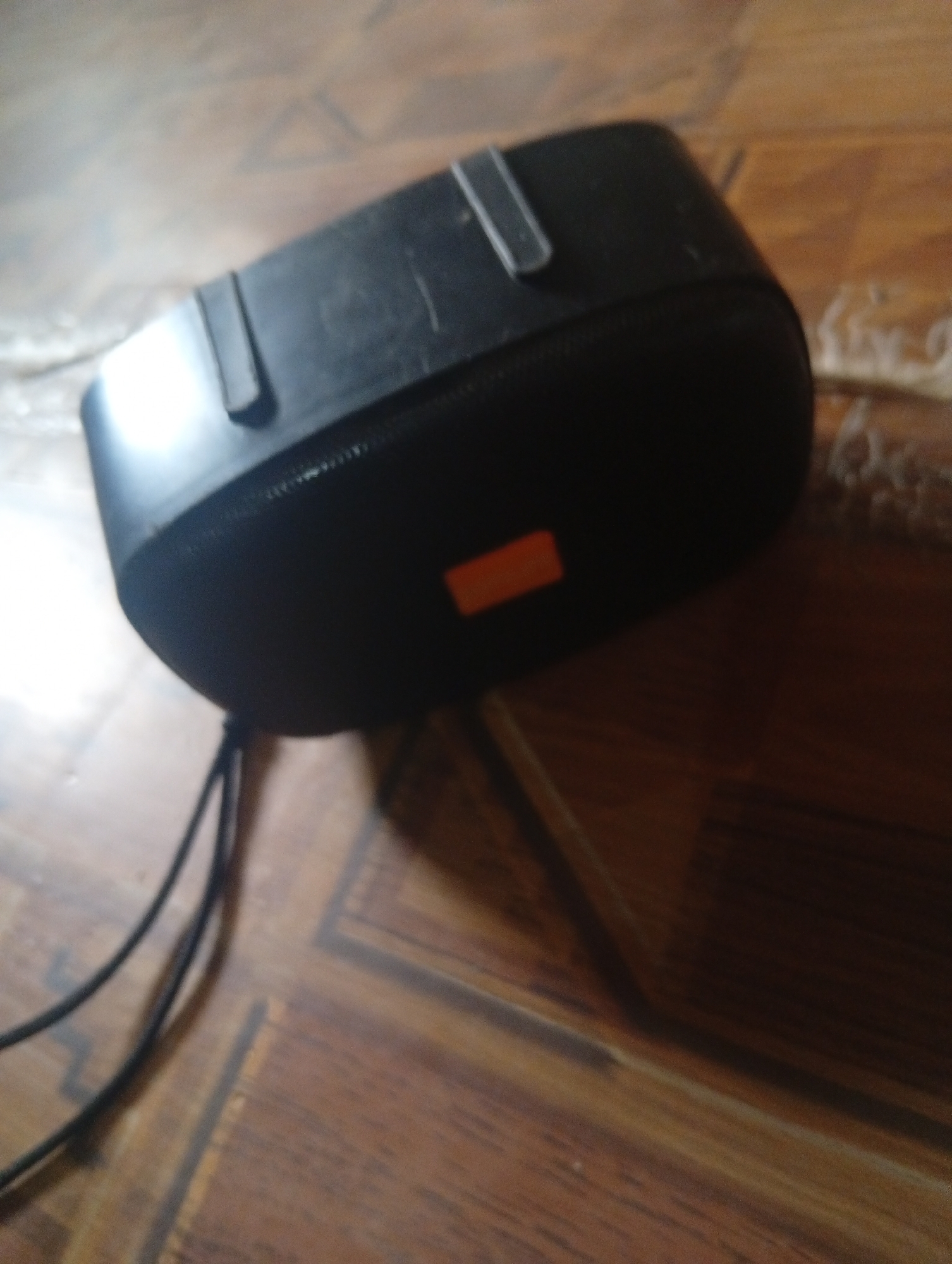 The image shows a black portable speaker with an orange rectangular logo on its surface, resting on a wooden floor with a visible wood grain pattern. Two black cables are connected to the speaker and extend out of the frame. The lighting suggests an indoor setting, possibly a room. There are no people or animals visible in the image. The scene is static and does not depict any action or interaction. The location in the city of Gulak, Nigeria, cannot be determined from the visual information provided in the image.
