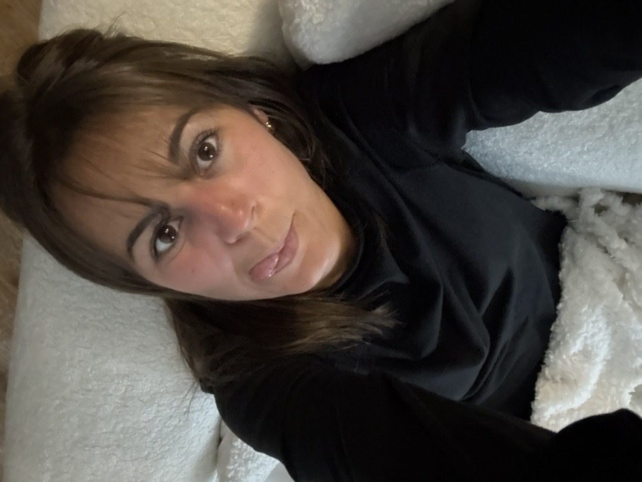 A woman with brown hair and bangs looks directly at the camera, her tongue slightly out. She's wearing a black long-sleeved shirt and lies on a white, fluffy surface, possibly a pillow or blanket. The camera angle is slightly tilted, giving a relaxed, casual vibe. She has fair skin with reddish cheeks, and visible eyelashes. Her expression suggests playfulness.