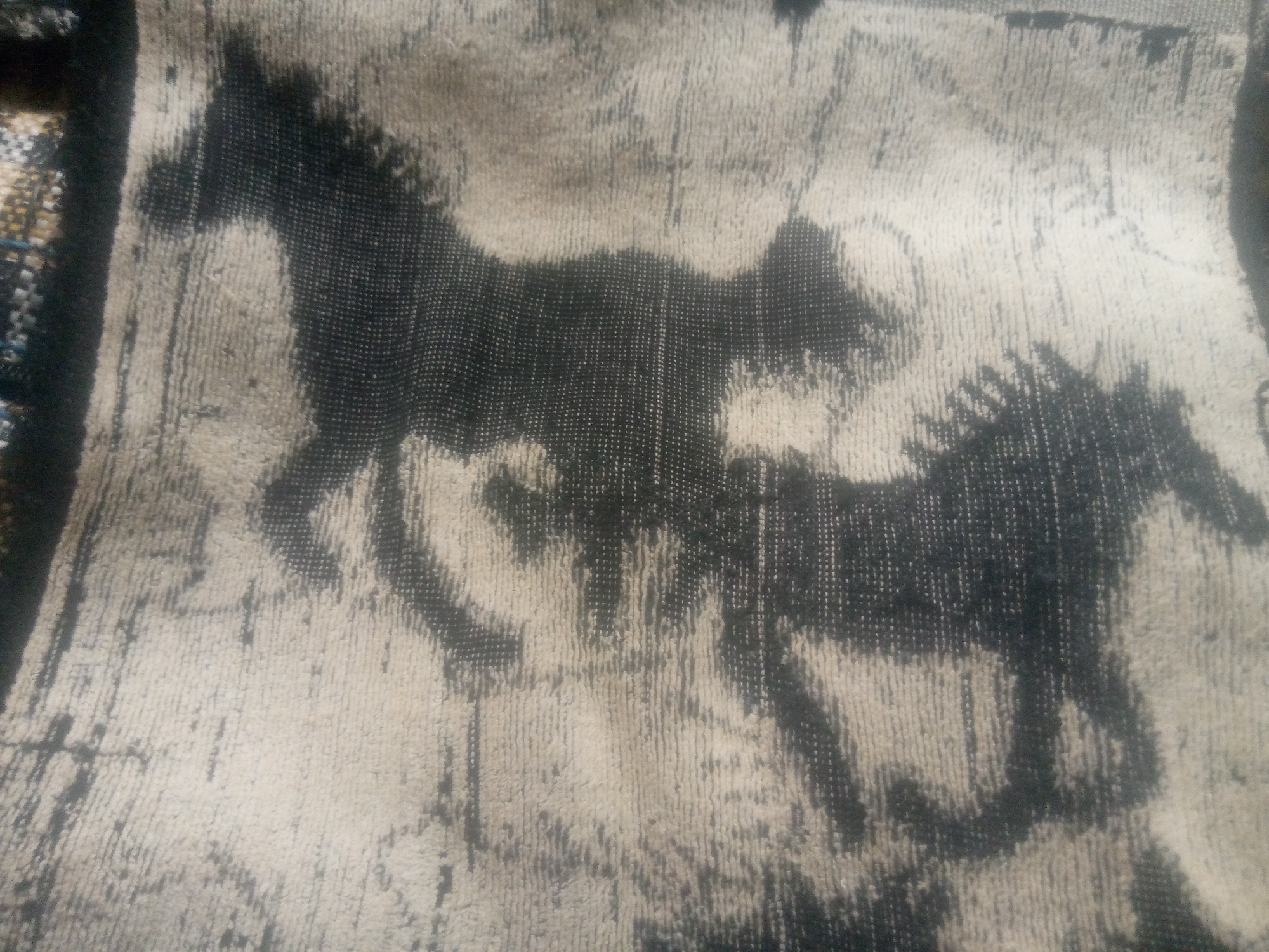 The image shows a close-up view of a textured fabric with a pattern of horses. The fabric appears to be a tapestry or rug, with a coarse weave creating a mottled effect. Two horses are depicted in motion, with their bodies rendered in dark, textured lines against a lighter, textured background. The horse on the left is more fully visible, with its mane and tail flowing. The horse on the right is partially obscured, but its hindquarters and legs are discernible. The overall color palette is muted, with shades of black, grey, and beige. The lighting is even, and there are no shadows or reflections to indicate a specific environment. The image is likely a photograph of an object, rather than a scene from a specific location.
