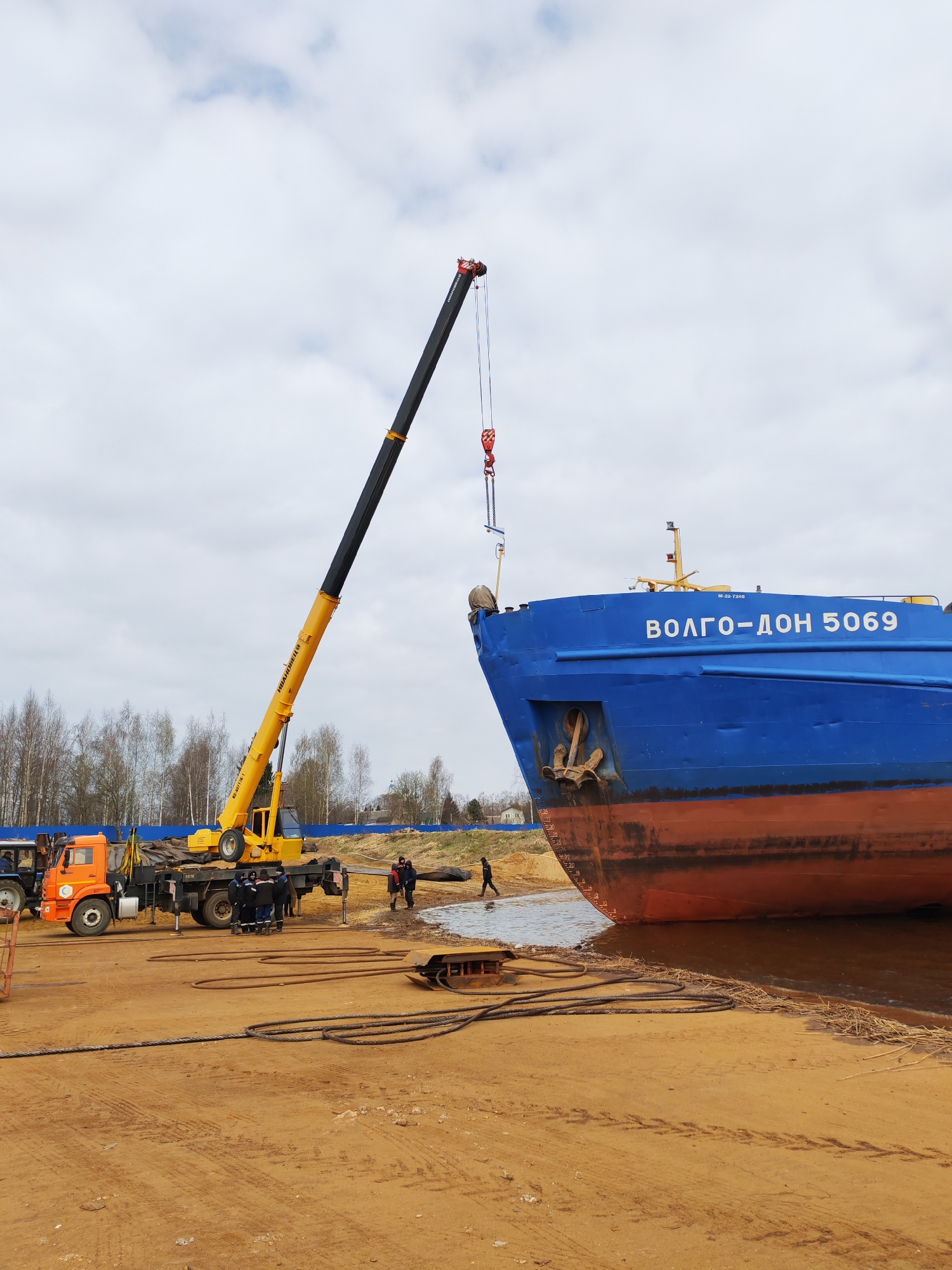 The image depicts an outdoor scene in Rybinsk, Russia, likely at a shipbuilding or repair yard. A large blue ship, identified as "ВОЛГО-ДОН 5069" (VOLGA-DON 5069) by the inscription on its bow, dominates the right side of the frame. The ship's hull is partially painted red below the waterline. A heavy-duty yellow crane with a black boom extends towards the ship's bow, suspending a metal component via cables and a hook.
Several people are present in the scene, including a small group standing near the base of the crane, seemingly observing the ongoing operation. Another person is walking near the waterline of a muddy area adjacent to the ship. Additional industrial vehicles, including an orange truck and a possible tractor, are parked to the left of the crane.
The ground is covered in sand and dirt, showing tracks and the marks of heavy machinery. Thick cables are coiled on the ground near the foreground. The background features a line of trees and a cloudy sky.