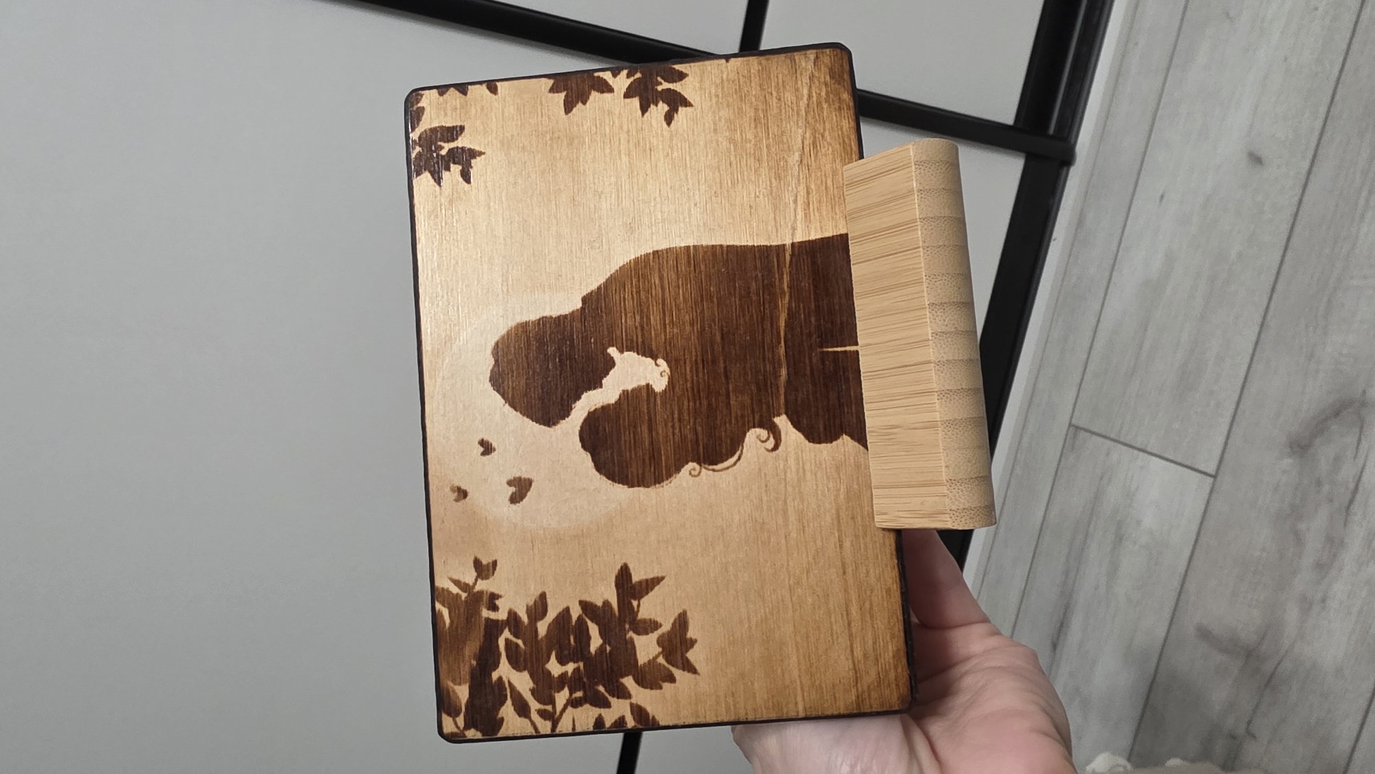 A close-up shot depicts a wooden bookstand featuring a burnt-in design. The design includes silhouetted figures resembling a person and a bear, foliage at the top and bottom, and heart-shaped figures. The stand itself has a bamboo-textured cylindrical prop attached to the back. The stand is held in a hand, likely indoors, given the background showing a wall and a section of a black metal frame. The location is Unknown, Unknown.