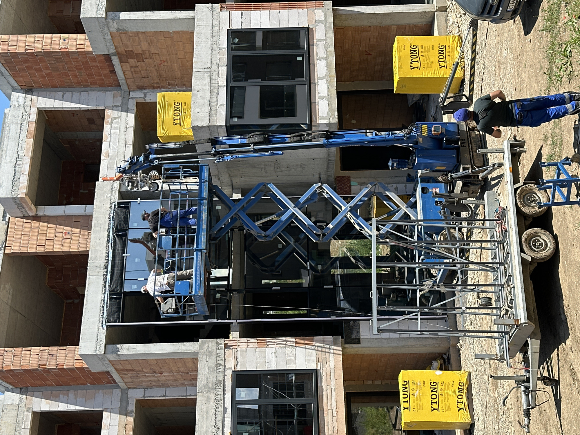 This image depicts a construction site in the city of Stara Zagora, Bulgaria, where workers are installing large glass panels on a multi-story building. A blue scissor lift is positioned against the building, with two workers on its platform actively engaged in the installation process. One worker, wearing a white cap and a light-colored shirt, is holding a tool. The other worker, in blue overalls, is also on the platform. Another worker, wearing a blue cap and blue work pants, is standing on the ground, looking towards the construction.
The building is under construction, with exposed brick and concrete elements visible. Several yellow packages with the "Ytong" logo are stacked on concrete ledges around the site. In the foreground, a metal trailer with a frame structure, likely for transporting glass, is parked. A dark-colored car is partially visible on the right side of the image. The ground is covered in dirt and gravel, typical of a construction environment. The sky is clear and the scene is illuminated by bright sunlight, suggesting it is daytime.