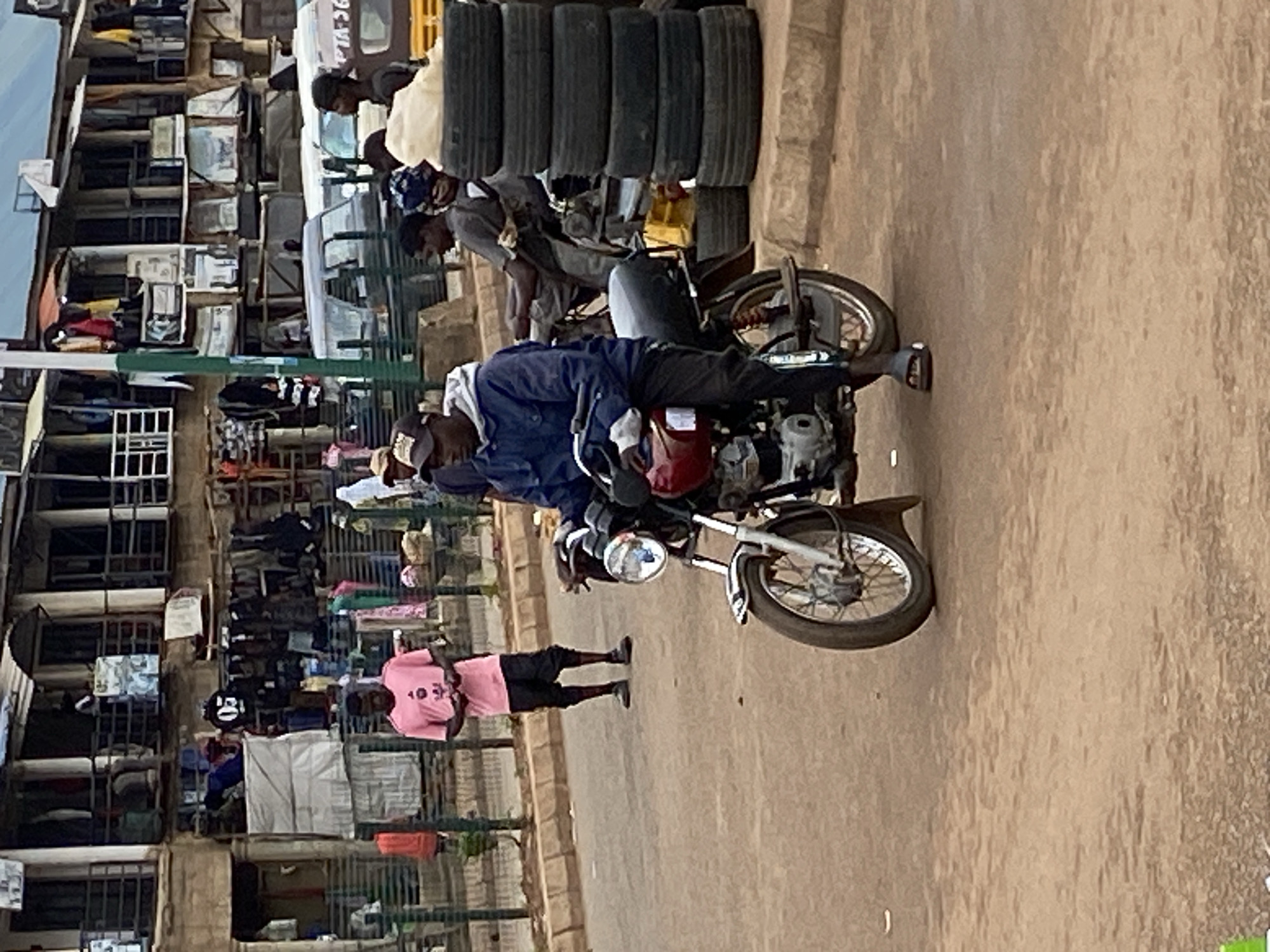 The image captures a street scene in Bukuru, Nigeria, likely a market area given the stalls and goods visible. In the foreground, a man wearing a blue jacket and dark pants rides a red motorcycle, facing right. His left foot is visible, wearing a dark sandal. Behind him, a stack of several black tires is piled on the right side of the frame. Further back, another man in a pink t-shirt and black shorts stands looking down, holding his hands together. He is positioned near a green metal barrier separating the sidewalk from the street. In the background, a two-story building with balconies and numerous stalls displaying clothing and other merchandise lines the street. Several vans and other vehicles are parked along the road. The overall environment suggests a bustling urban area with commercial activity.