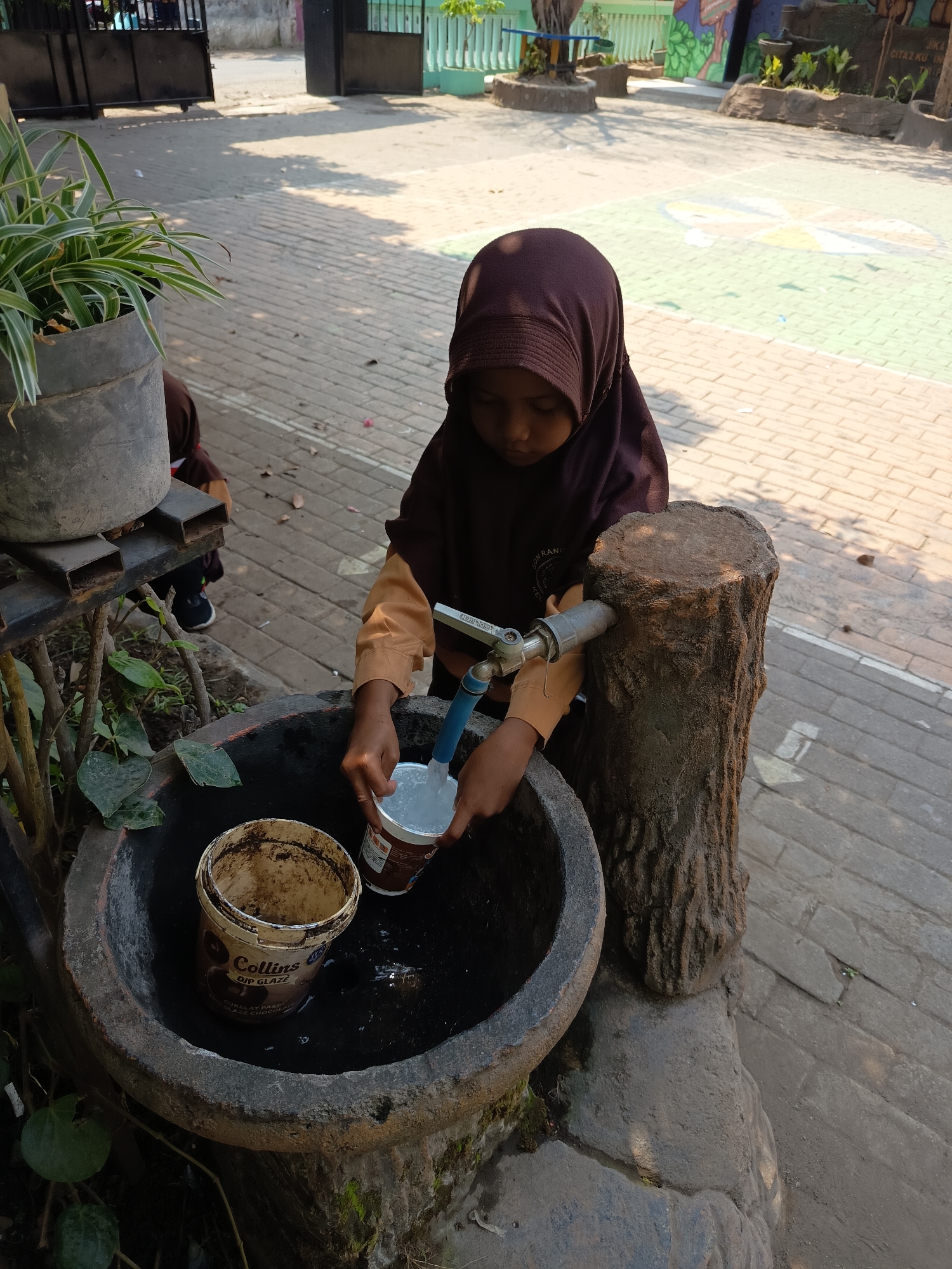 In Widoro, Indonesia, a young girl wearing a brown hijab and school uniform is filling a plastic cup with water from a spigot. The spigot is attached to a pipe that protrudes from a decorative tree stump. The girl is standing next to a large, weathered concrete basin, which is partially filled with murky water. Inside the basin, a dirty white bucket with a "Collins Dip Glaze" label is visible. To the left of the basin, a plant in a gray pot sits on a metal stand. The ground is paved with light-colored bricks, and a shadow of the girl and the basin is cast on the ground, indicating a sunny day. In the background, a black metal gate and a colorful mural on a building are visible. Another child in a similar uniform is partially visible in the background, walking away from the scene.