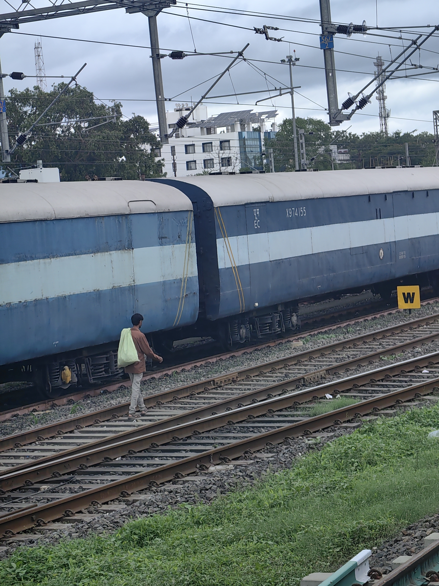 In Khandwa, India, a man walks along railway tracks beside a blue and white passenger train. He carries a light green bag over his left shoulder. The train has the number X974155 printed on its side. Overhead, a complex network of electrical wires and poles supports the train's power supply. In the background, trees and modern buildings are visible under a cloudy sky. The railway tracks are surrounded by gravel and green vegetation.