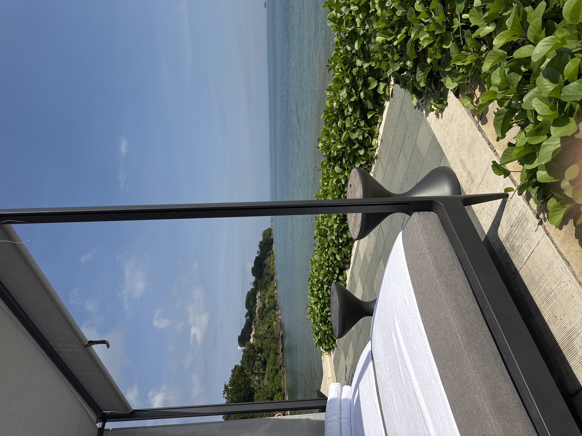 The image presents a partial view of a coastal scene, potentially taken from a patio or balcony setting in Kuta Selatan, Indonesia. The ocean and an island covered in lush vegetation are visible. There is a structure, partially in frame, with a grey canopy and metal frame, suggesting it may be part of a seating area. A chaise lounge with white towels on it sits next to two black hourglass-shaped end tables. Adjacent to the lounge, green foliage is visible. The scene is captured on a sunny day with blue sky.
