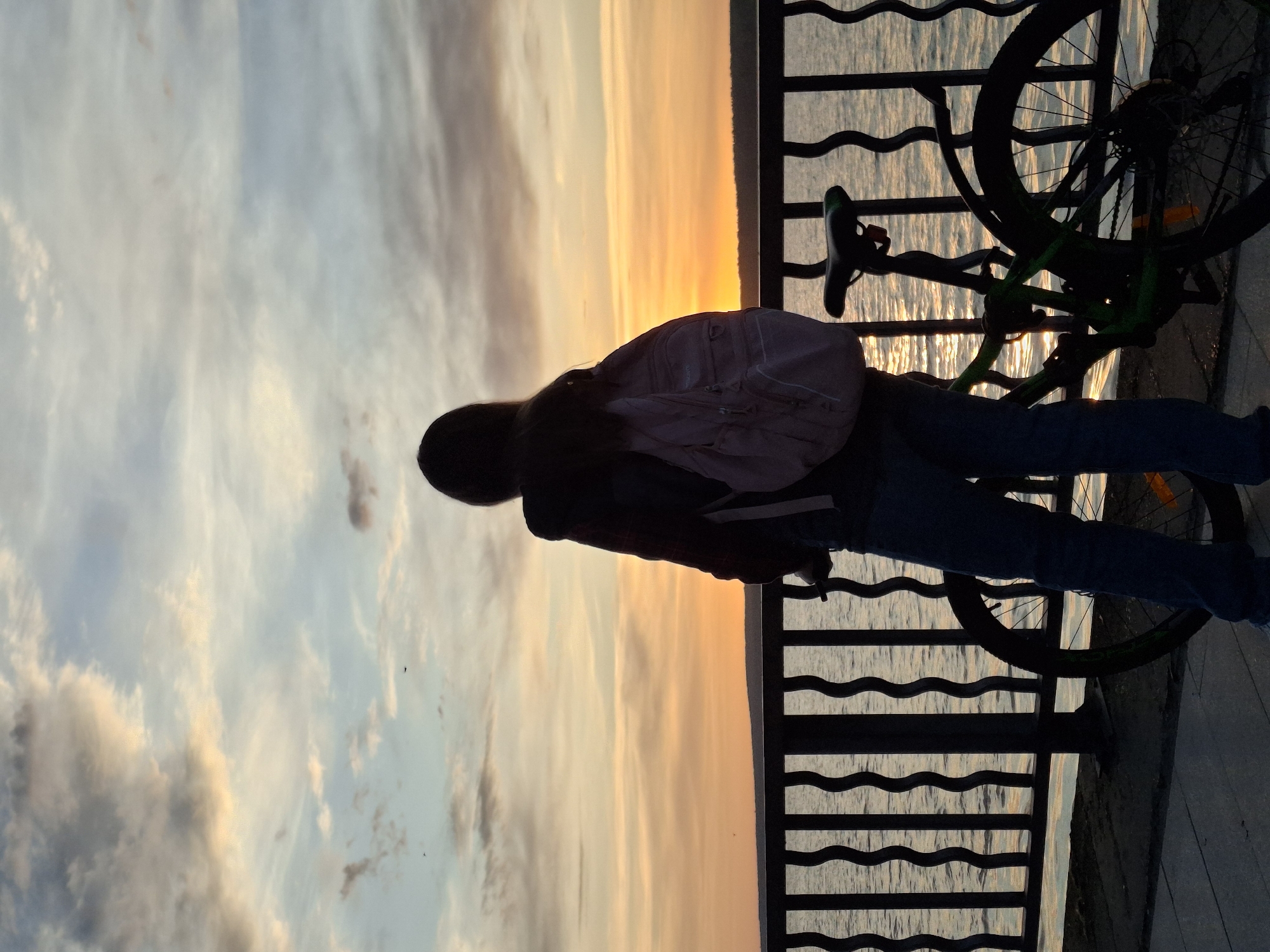 A person stands at a railing in the city of Chaikovsky, Russia. The subject is wearing blue jeans, a dark jacket, and a light pink backpack. A bicycle is propped against the railing to the right. The railing overlooks water. The sky shows shades of blue and orange, suggesting either sunrise or sunset.