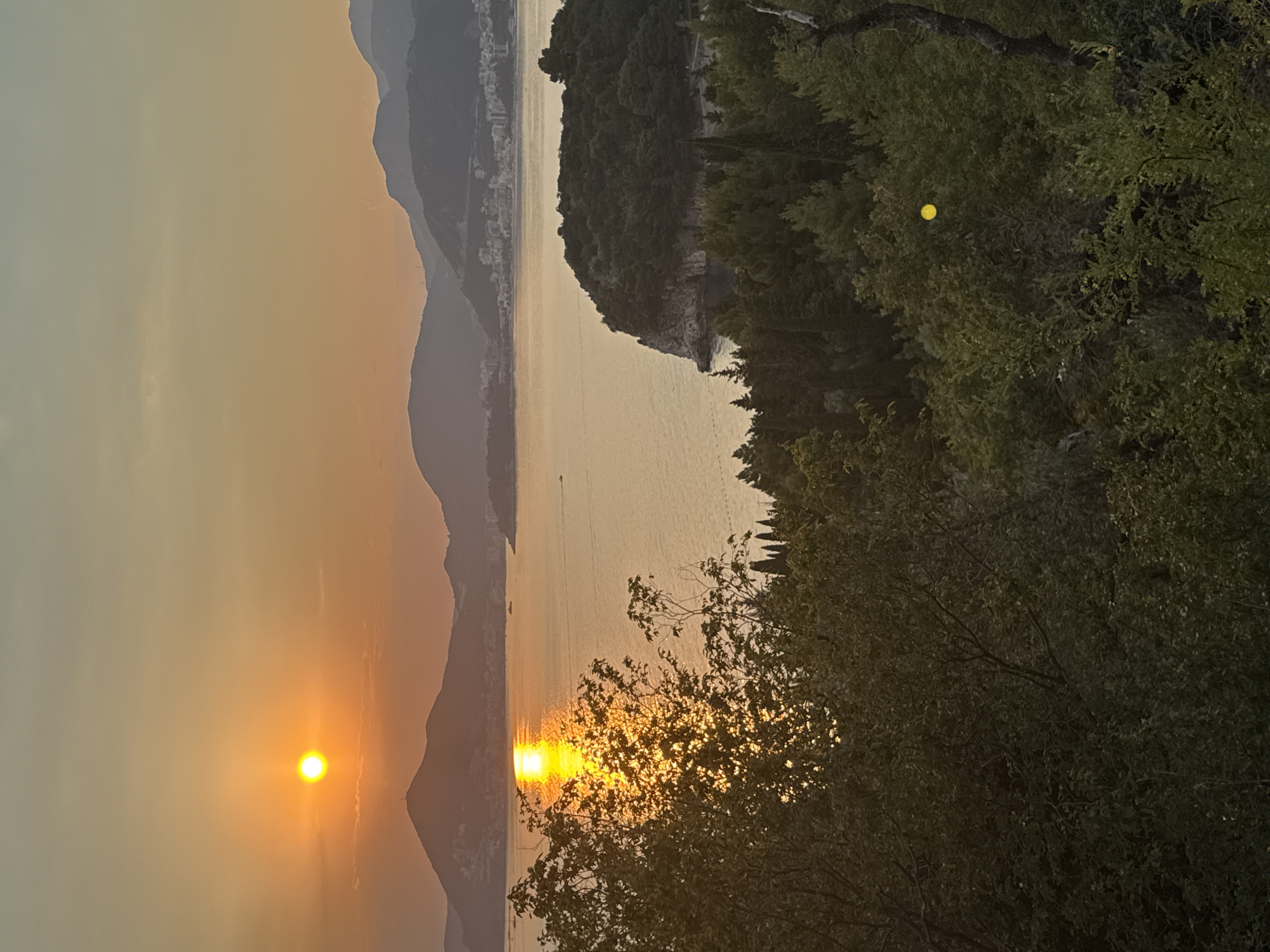 This is a photograph taken at sunset, possibly on a hazy or overcast day, casting a warm, orange glow across the scene. The image is oriented vertically.
The main subjects include a body of water, likely the sea, reflecting the sunlight. In the distance, silhouetted mountains rise, some with buildings faintly visible on their slopes, suggesting a coastal town or resort. A large, tree-covered island or peninsula juts out into the water from the right side of the frame. The foreground is dominated by the dense foliage of trees and branches, with dappled sunlight filtering through. A small, bright yellow object, possibly a lantern or a decorative element, is suspended among the leaves of the trees on the right. The sun, a brilliant orb of orange, is low in the sky, situated in the left portion of the frame, creating a strong reflection on the water. The overall mood is serene and tranquil, characteristic of a sunset. No visible text is present in the image. The location context suggests this scene is likely Sveti Stefan, Montenegro.