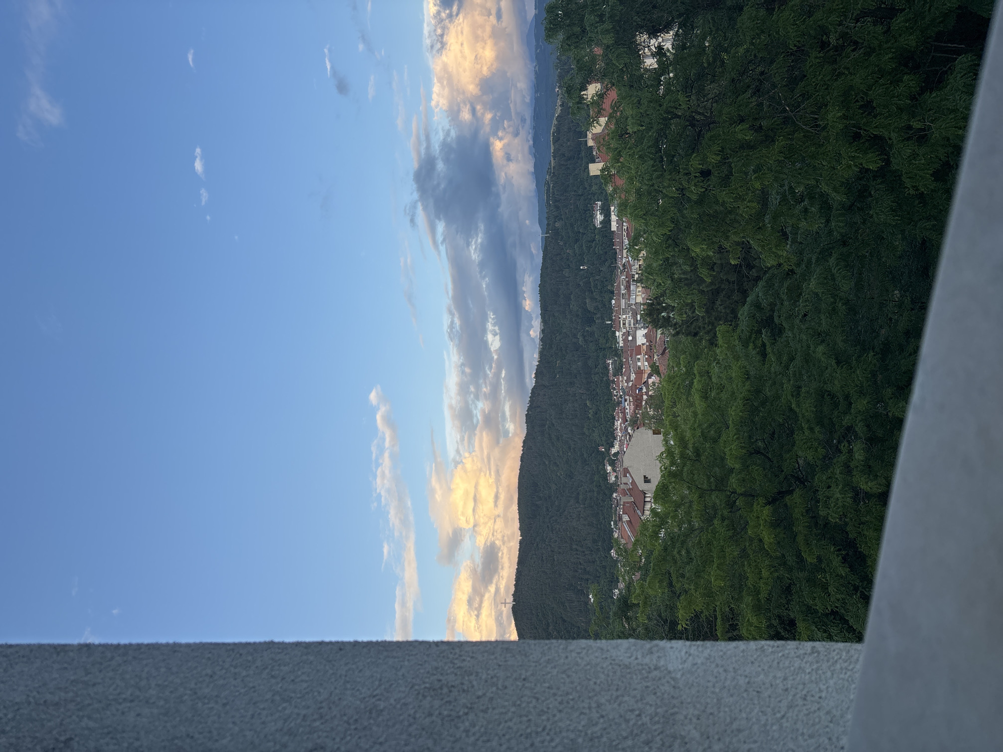 This image, likely captured from a balcony or elevated viewpoint in Blagoevgrad, Bulgaria, presents a serene, expansive vista of a town nestled within a mountainous landscape during late afternoon or early evening.
In the foreground, a textured, grey concrete wall or railing forms the base of the frame, with a lighter, vertical structure on the right side. Lush, dark green trees fill the immediate midground, their dense foliage obscuring parts of the town below.
Beyond the trees, a charming town unfolds, characterized by numerous buildings with distinctive reddish-brown tiled roofs and light-colored facades. These structures are closely packed, indicating an urban or residential area. The town stretches across the base of a majestic, densely forested mountain that dominates the central midground, its peaks forming a prominent horizontal line across the frame.
The background features a broad expanse of clear blue sky, dotted with scattered clouds. Some clouds glow with warm golden-orange and soft pink hues, especially those closer to the mountains, strongly suggesting the golden hour approaching sunset. Other clouds are a more neutral grey. In the far distance, beyond the immediate forested mountain, fainter, lighter blue mountain ranges are visible on the horizon. A small, white object resembling an airplane can be seen in the sky above the forested mountain.
The overall scene conveys a tranquil atmosphere, showcasing the natural beauty of the region with its interplay of urban development and dramatic mountain scenery under a beautiful evening sky. There is no visible text or identifiable people in the image.