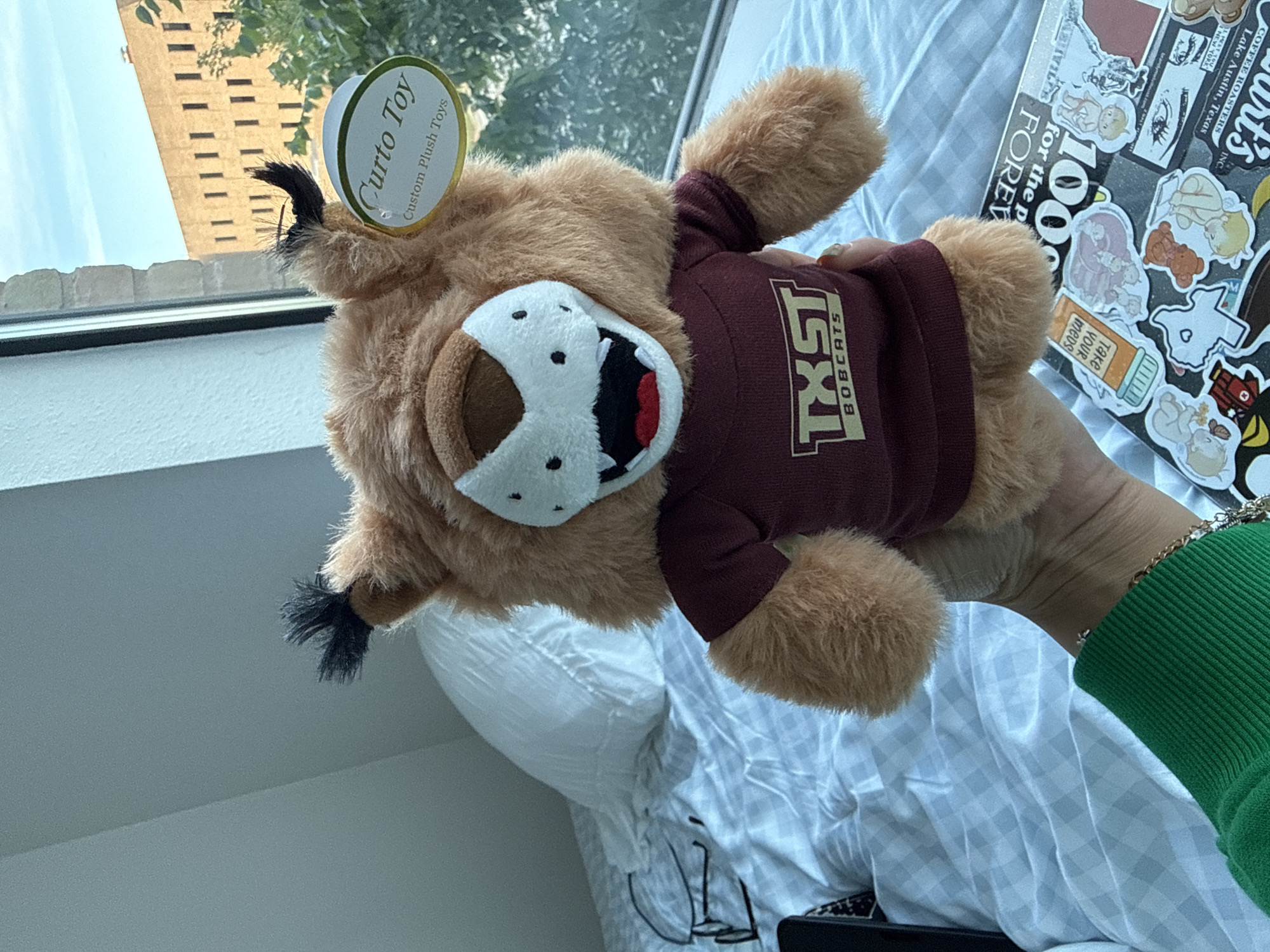 A bobcat plush toy, wearing a maroon shirt printed with "TXST Bobcats," is displayed against a backdrop of a window and the outside world. The toy is held in a person's hand, with a green sleeve visible. The location is identified as Austin, Texas, based on stickers on a laptop to the right of the toy. In the background, a tall brick building is partially visible through the window. The plush toy has a tag labeled "Curto Toy Custom Plush Toys" attached to it.