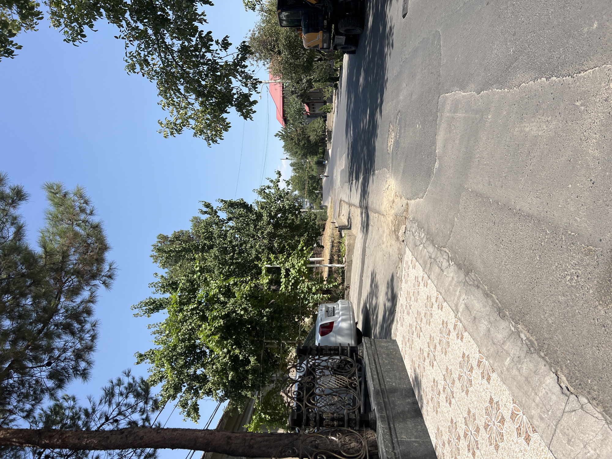 A sunny, clear day illuminates a quiet, tree-lined residential street in an unspecified urban location. A paved road, showing signs of repair and some cracking, stretches into the distance, bordered by sidewalks and residential properties. On the left, a decorative metal fence and a tiled walkway front a property. A white sedan is parked on the side of the road. Lush green trees and foliage line both sides of the street, casting dappled shadows on the pavement. In the background, a red-roofed house is visible. On the right side of the road, a yellow construction vehicle with large tires is parked. A lone cyclist is visible in the distance, riding down the street. The overall scene suggests a peaceful, suburban environment.