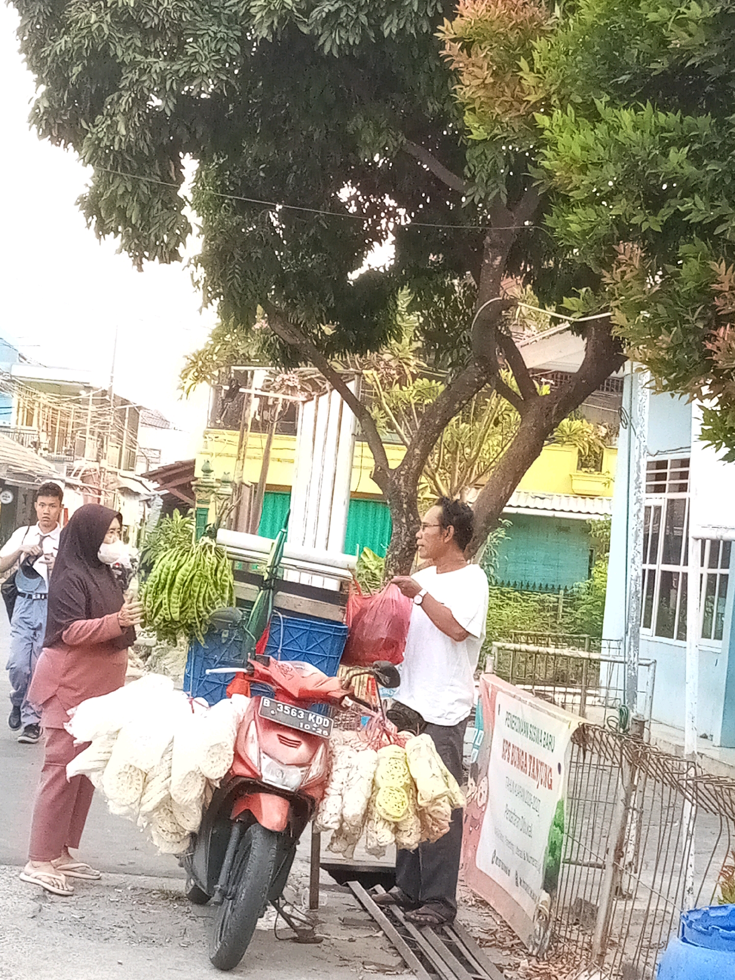 The image depicts a street scene in Bekasi, Indonesia, with a focus on vendors and their goods. In the foreground, a red motorcycle is laden with large quantities of what appear to be puffed rice snacks. To the left of the motorcycle, a woman wearing a dark hijab and a pink outfit is seen interacting with the snacks, while a young man in a white shirt and blue pants walks behind her. To the right of the motorcycle, a man in a white t-shirt holds a red plastic bag and stands next to a cart displaying more of the puffed rice snacks, along with some green produce which resembles petai beans. A banner with Indonesian text is visible behind the man. The background shows a street with buildings, trees, and fences. The environment suggests a commercial or market area within the city.