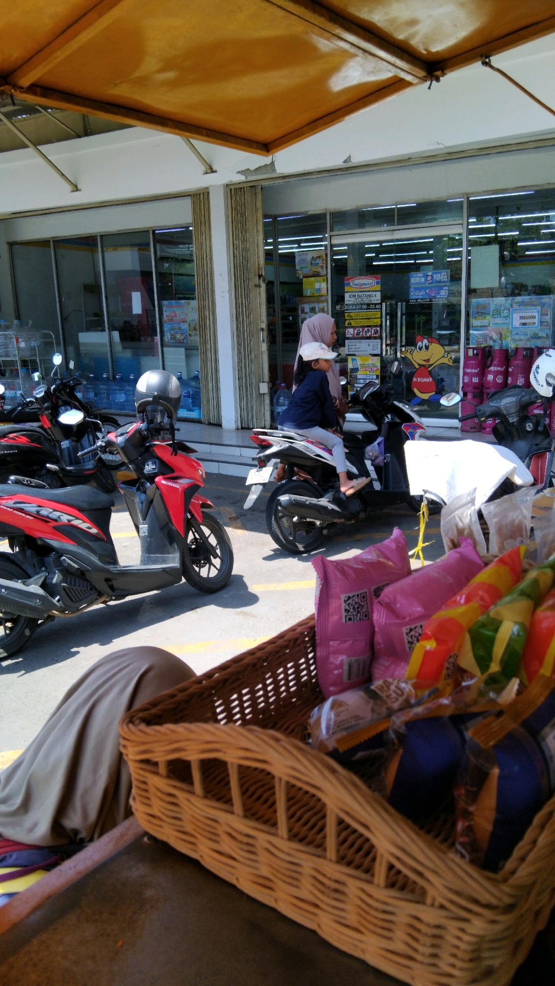 The image shows a street scene in front of a convenience store, likely in Watesalit, Indonesia. In the foreground, a wicker basket is filled with packaged snacks and drinks. Behind the basket, several motorcycles are parked. One red motorcycle is prominently displayed. Further back, a woman wearing a headscarf and a child in a hat are seated on a white and black motorcycle. The convenience store has large glass windows revealing shelves stocked with products. The building has a yellow awning extending over the storefront. The ground is paved, with yellow lines indicating parking spaces. The lighting suggests it is daytime, with sunlight casting shadows.