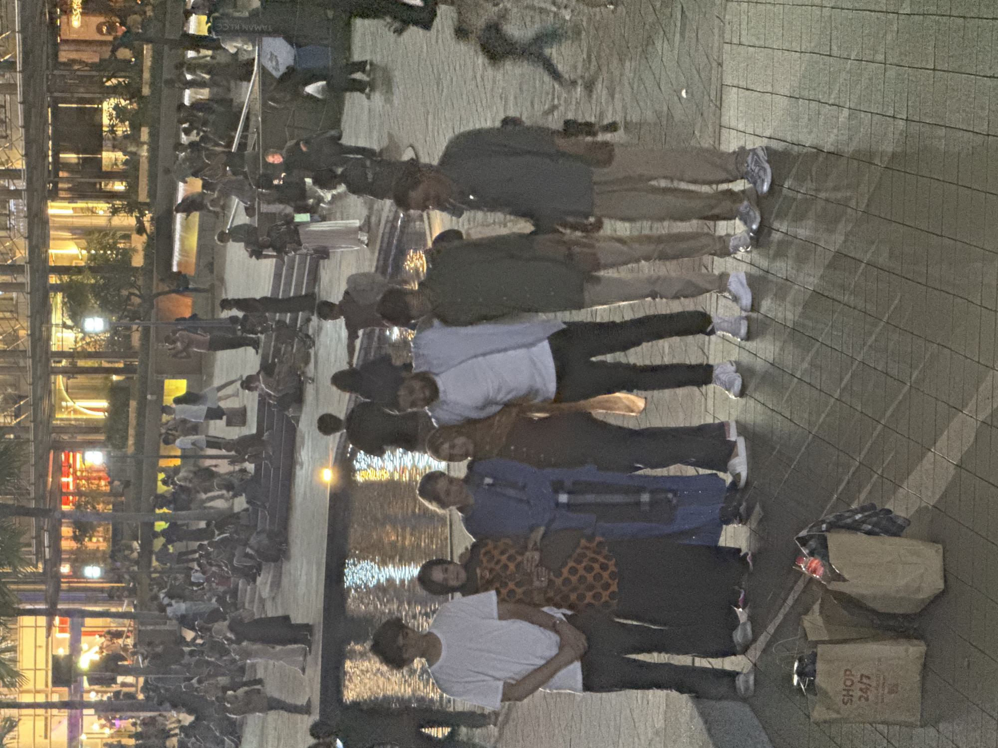 The image depicts a nighttime scene in a bustling public square or park in Kuala Lumpur, Malaysia. In the foreground, a group of seven individuals is standing, posed for a photograph, generally facing the camera. They are casually dressed in t-shirts, shirts, and trousers, appearing relaxed with some smiling.
Behind this primary group, a large body of water, likely a fountain or decorative pond, reflects the bright artificial lights from surrounding buildings, creating striking patterns on its surface. Numerous other people are visible in the background, some standing and others seated on steps or ledges around the water feature, suggesting a gathering or general evening leisure activity. Modern buildings with brightly lit windows form the backdrop, indicating an urban setting.
The ground is paved with light-colored tiles. To the right of the foreground group, two brown paper shopping bags are placed on the ground; one is partially open, and the other clearly shows the text "SHOP 24/7." In the distant background, faint text "TAMAN KLCC" can be discerned, confirming the location as KLCC Park. The scene is illuminated by artificial city lights, giving it a vibrant, yet intimate, evening ambiance.