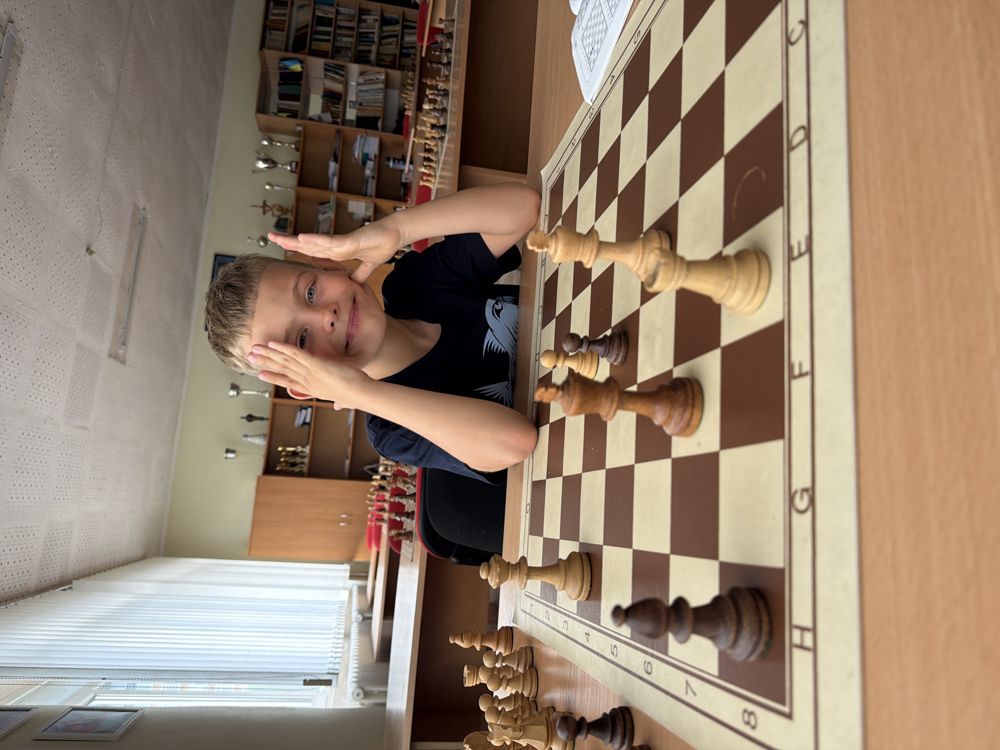A young boy with blonde hair and blue eyes is sitting at a wooden table, looking at the camera with a playful expression. He is wearing a dark blue t-shirt and has his hands resting on his cheeks. In front of him is a chessboard with various chess pieces set up, some in play and others lined up. The chessboard is placed on a wooden desk. In the background, there are bookshelves filled with books, trophies on shelves, and a window with white blinds. The room appears to be a classroom or a library. The lighting is natural and comes from the window.