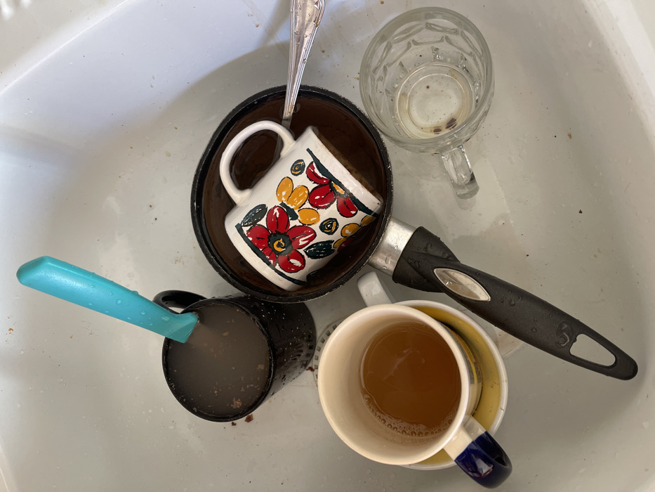 An overhead shot captures a collection of dirty dishes accumulated in a kitchen sink in Dosso (Ciappanico), Italy. The sink, predominantly white, shows visible grime, stains, and food particles on its surface.

The subjects are various unwashed kitchen items:
- A dark cooking pot, possibly made of coconut shell or a similar material, contains a white ceramic mug adorned with red, orange, and yellow floral patterns and green leaves. A silver spoon rests inside this mug.
- To the upper right, a clear glass mug or stein with a textured, patterned surface is mostly empty, containing some dried residue at the bottom.
- Towards the lower left, a black ceramic mug holds a light blue handled utensil, resembling a spatula or a small knife, which is partially submerged in a dark, murky liquid, likely containing coffee grounds or similar residue.
- At the bottom right, a white ceramic mug with a yellow interior and a blue stripe on its handle contains a light brown liquid, consistent with leftover tea. This mug appears to be stacked on top of another dish or mug.
- The dark cooking pot also features a black handle with silver accents, with water droplets visible on its surface and on the blue utensil handle, suggesting recent use or rinsing.

The scene depicts the aftermath of a meal or beverage consumption, with dishes left unwashed in the sink. No people are present, and no specific activity is actively occurring beyond the passive state of waiting to be cleaned. There is no visible text in the image, and no specific time of day or weather conditions can be inferred.