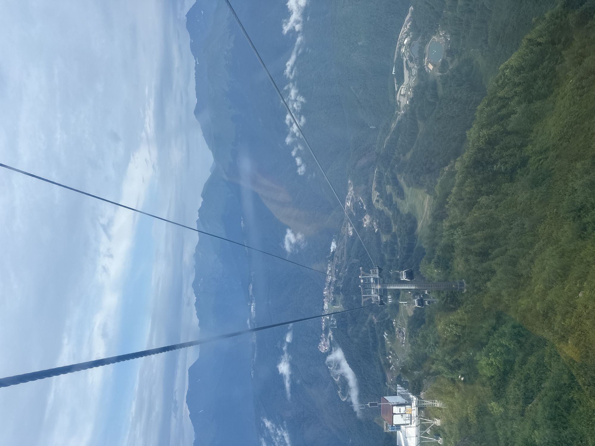 The image captures a scenic view from a cable car traveling over the mountainous terrain near Estosadok, Russia. In the foreground, thick green forest covers the slopes, transitioning to a more distant view of mountains shrouded in mist and clouds. A cable car station with a white building and a metallic structure is visible on the left. Several cable cars are suspended from cables, with one prominently featured in the middle right of the frame, descending a ski slope. Further down the valley, a cluster of buildings, likely a resort or village, is nestled amidst the greenery. The sky is overcast with scattered clouds.