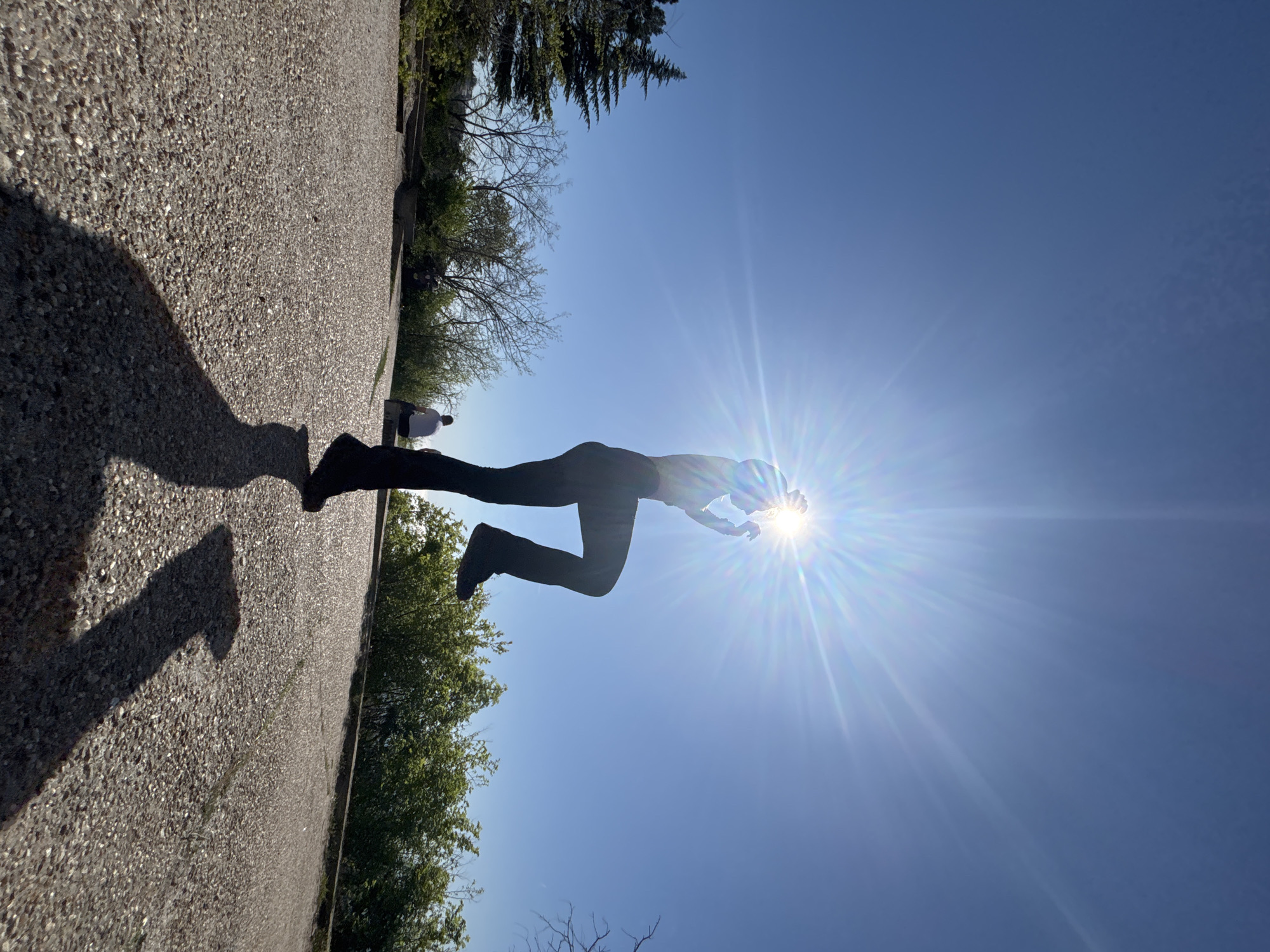 A person, likely an adult, wearing dark clothing (possibly leggings and a long-sleeved shirt) is silhouetted against a bright sun in a clear blue sky. The sun's glare obscures their features. The person appears to be in mid-air, possibly jumping or posing. A shadow cast by the person is visible on a textured ground surface (possibly asphalt or a paved path) to the left of the frame. The background includes trees and greenery, suggesting a park or outdoor setting. Another person is walking in the distance. The photograph is taken from a low angle and is oriented with the sky above and the ground to the left. The location is Unknown, Unknown.