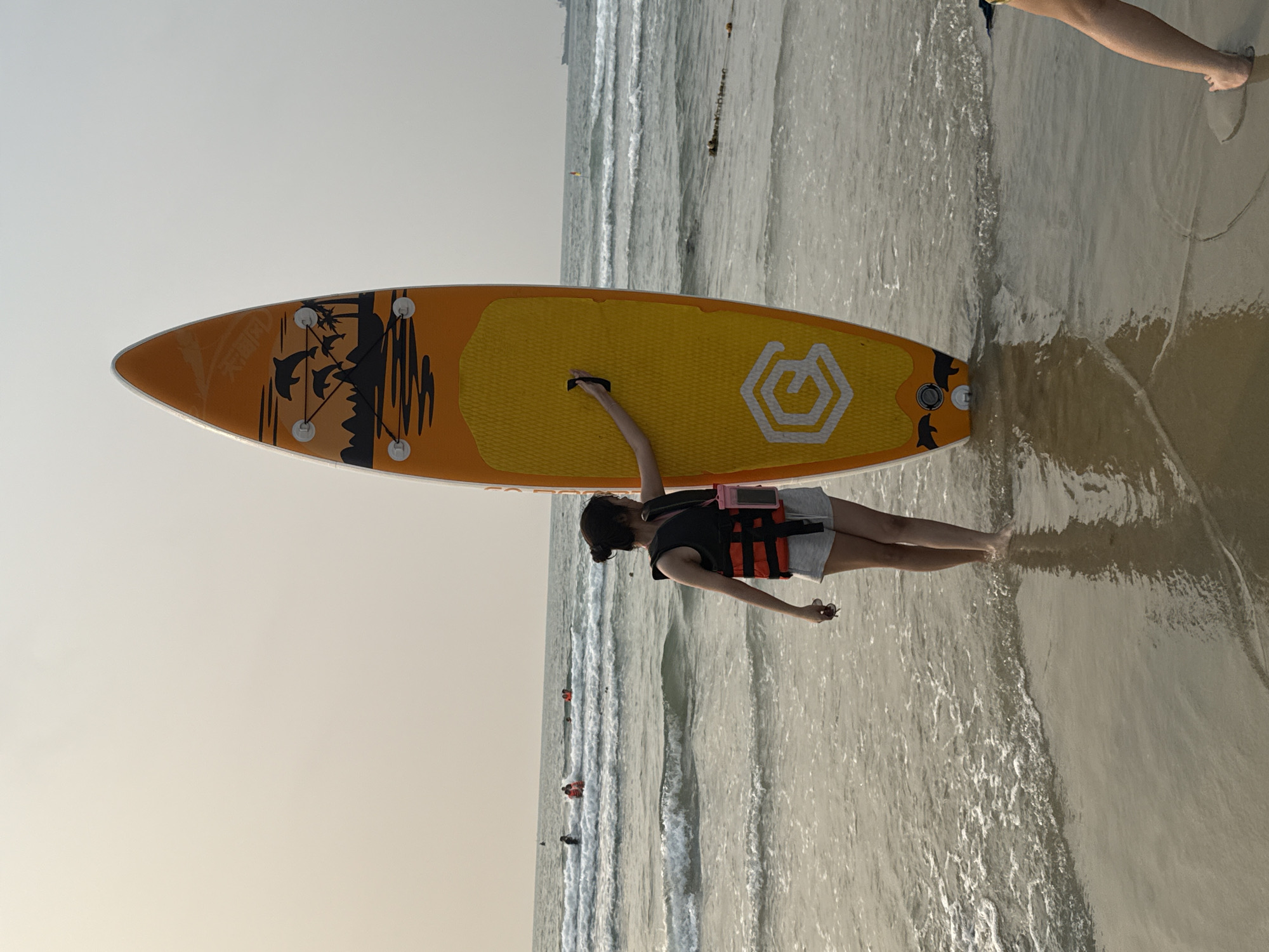 A person is lying horizontally on a beach next to a standup paddleboard. The person is wearing a black shirt, gray shorts, and a red life vest. The paddleboard is yellow with an orange perimeter and has a geometric white logo on the yellow area. The person is lying on the edge of the water, with waves washing around them and the paddleboard. Other people are visible in the water farther out in the ocean. The sky is overcast, suggesting either early morning or late afternoon. This is likely Mỹ Khê Beach in Đà Nẵng, Vietnam.