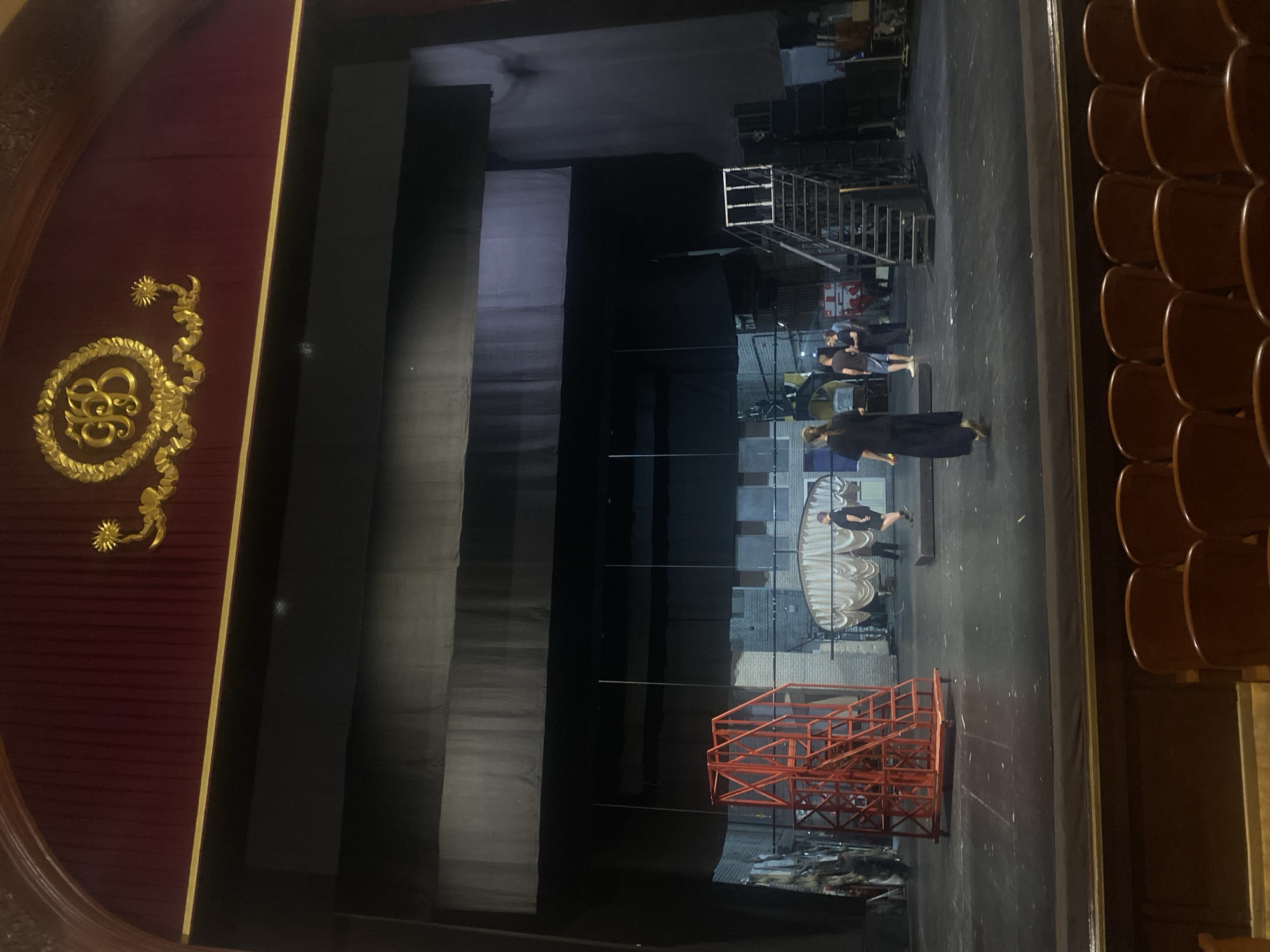 The image captures the interior of what appears to be a theater or performance venue in Шебунино, Russia. The shot is taken from the audience seating area, showing rows of empty wooden chairs on the right side. To the left, a section of the stage is visible, with a red and gold ornamented wall bordering the stage's edge. The stage itself has several people working on set construction or maintenance. There are scaffolding structures, pieces of set design, and what looks like metal framing on the stage floor. People are seen near the stage, some standing and seemingly conferring, others potentially working on the set. The environment suggests preparations for a theatrical production or event.