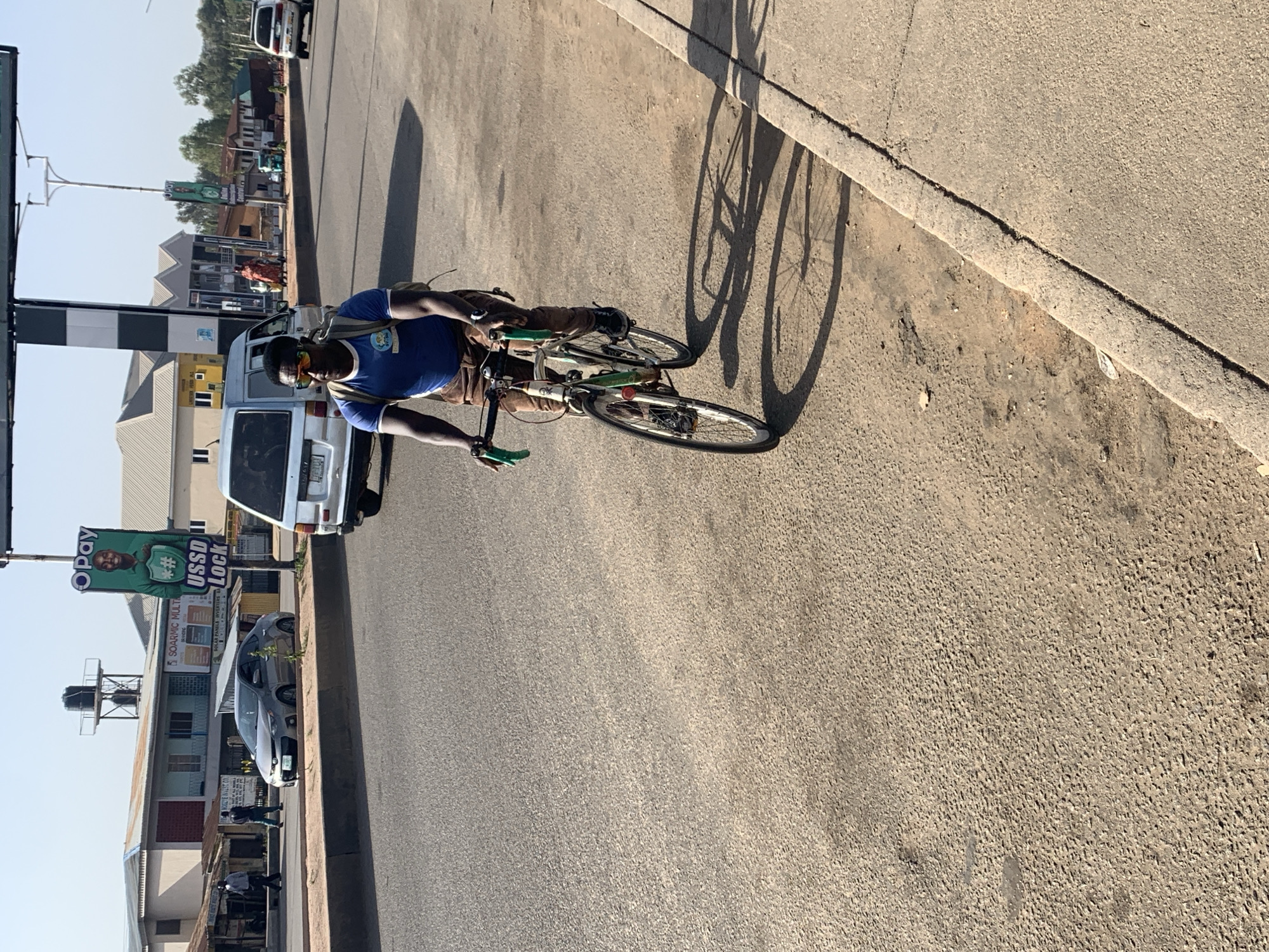 A man on a bicycle rides on a paved road in Jos, Nigeria. The man is wearing a blue t-shirt, sunglasses, and a backpack. The bicycle is green and has panniers. The street has a curb and sidewalk on the right. A sign for "Opay USSD Lock" is visible on the left, as are buildings and parked cars. The weather appears sunny.