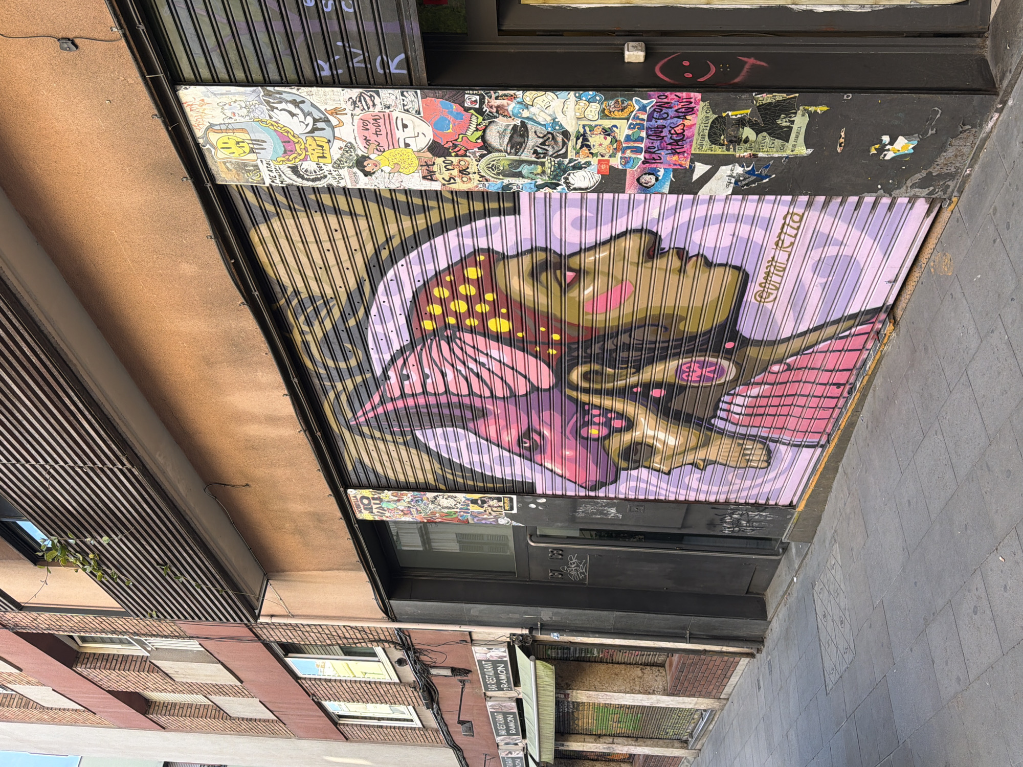 This is a street-level, medium-angle outdoor photograph of a narrow city alleyway in Barcelona, Spain. The perspective is from the sidewalk, looking down the alley. The predominant elements are the facades of buildings on the right and left, featuring rolling metal shutters that are mostly closed.
On the left side of the alley, there are two commercial establishments with green awnings. The closest one has a sign reading "BAR RESTAURANT RAMON." Its rolling shutter is partially pulled up, revealing a display inside. Further down the alley on the left, there's another establishment with a similar awning and a closed shutter. The building on the left is constructed with red brick and beige concrete accents.
The focus of the image is the large, colorful mural painted on the rolling shutter of a building on the right side of the alley. The mural depicts a stylized profile of a person's head, rendered in shades of pink, yellow, and purple. The head appears to be in profile, facing right, with a prominent headdress or hairstyle featuring yellow dots. There is also a superimposed image of a skull with animalistic ears or features in a pinkish hue. Below the main mural, the artist's tag "@omr_ietza" is visible. To the right of the mural, there is a vertical column plastered with numerous small stickers and posters featuring various cartoonish characters, skulls, and text in Spanish. One sticker has a smiley face with toast for a head.
The alleyway itself has a paved sidewalk made of dark grey square tiles. A square metal grate is embedded in the sidewalk in the foreground. The lighting suggests it is daytime, with sunlight casting shadows on the sidewalk. The overall impression is one of an urban environment decorated with vibrant street art.