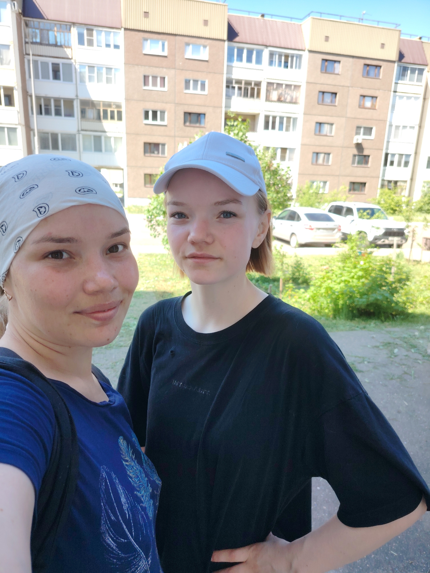 Two young women stand outdoors in front of a multi-story apartment building. The woman on the left wears a blue t-shirt with a feather design and a white bandana. The woman on the right wears a black t-shirt and a white baseball cap. In the background, cars are parked near green vegetation. The location is Марковский, Russia, as indicated by the buildings.