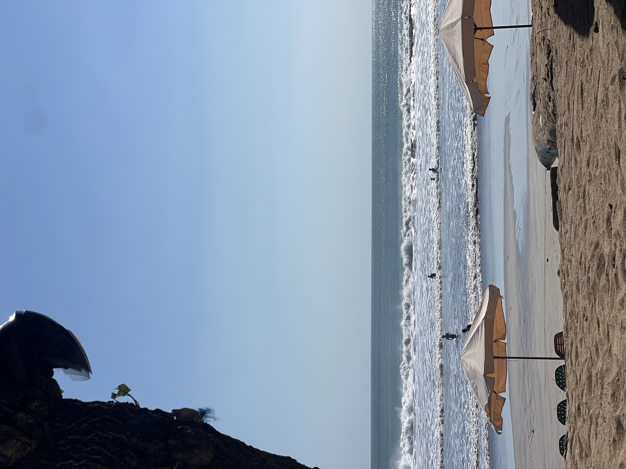 The image captures a serene beach scene in Kuta, Indonesia, on a clear, sunny day. The foreground is dominated by sand, with two large, light-colored beach umbrellas casting shadows. Several empty beach chairs are visible behind the umbrellas. In the middle ground, the gentle waves of the ocean roll onto the shore, with several people visible in the water. Some are standing, while others appear to be swimming or wading. The ocean stretches out to the horizon, meeting a clear blue sky. A large, dark object, possibly a tree trunk or a rock formation, is partially visible in the upper left corner of the frame, adding a natural element to the scene. The overall atmosphere is one of tranquility and relaxation.