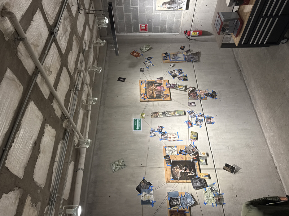 The image depicts an interior space in León, Mexico, characterized by an industrial aesthetic. The ceiling is unfinished concrete with rectangular recessed panels and exposed electrical conduits, from which multiple track lighting fixtures and a linear pendant light are suspended. The left wall is smooth gray concrete, hosting a large, intricate display. This display features numerous small photographs and printed images, secured with blue painter's tape and interconnected by a web of thin dark strings, some attached to three larger wooden panels within the arrangement. A green sign with white text reading "RUTA DE EVACUACION" (Evacuation Route) and an arrow points left above the display. The right wall is constructed from gray concrete blocks and features a red fire extinguisher mounted near the corner. A framed artwork, depicting a monochrome portrait, hangs further along this wall. In the lower right foreground, a black mobile utility cart with multiple drawers and a light-colored wooden top holds a red and silver espresso machine. Part of a dark wooden table extends from the right, behind the utility cart. The lower left corner shows the partial back of a person wearing a dark olive green long-sleeved shirt. The floor is smooth, unpainted concrete.