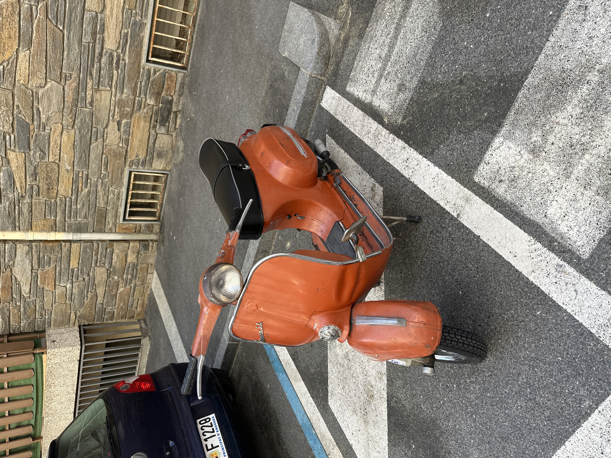 The image, taken in Encamp, Andorra, depicts a vintage, rust-orange Vespa-style scooter parked on an asphalt surface during daylight. The scooter, which shows signs of wear, features a black seat with white piping, chrome trim, and a prominent headlight. Faint text, including "Vespa" and "75 p", is visible on its body.
To the left of the scooter, the rear portion of a dark blue car, likely a hatchback, is visible. Its license plate clearly displays "PRINCIPAT D'ANDORRA," "F 1228," and the European Union flag with the country code "AD," confirming the location as Andorra. Partially visible next to the car is a wooden slatted structure.
In the background, a building with a textured stone facade rises, featuring two small, barred windows. The ground is dark asphalt, marked with white and a blue painted line, suggesting a designated parking area or street. The scene is brightly lit by the sun, casting distinct shadows from the scooter and other elements onto the pavement. No people are present, and the overall impression is that of a quiet, parked street scene.