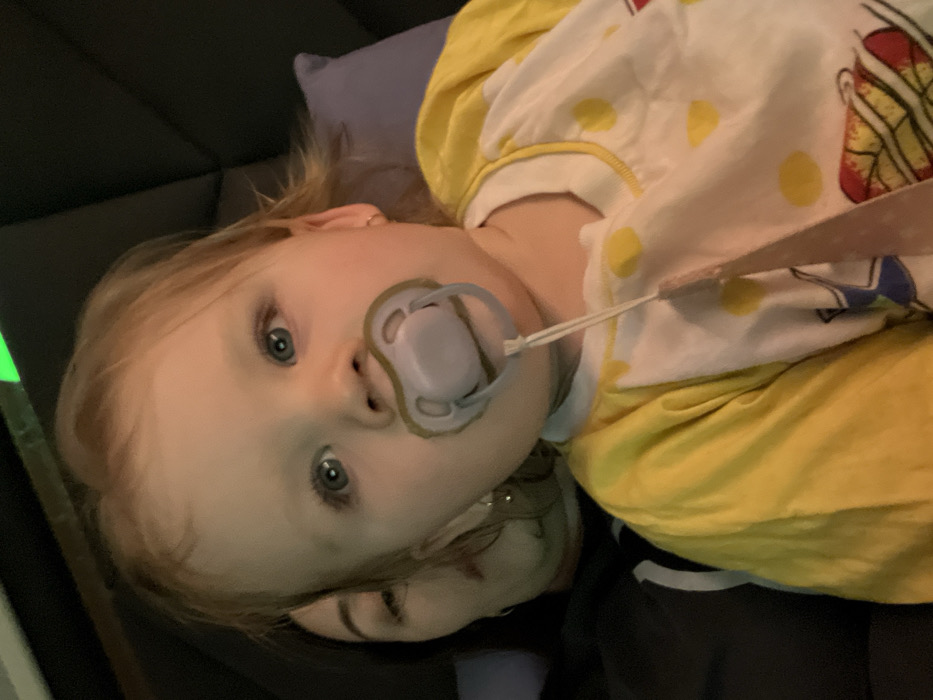 A close-up, frontal image depicts a toddler and a partially visible adult in an indoor setting. The toddler, positioned centrally, has light hair, blue eyes, and is looking directly at the camera. A light purple pacifier with a white string is in the child's mouth. The child wears a long-sleeved shirt that is yellow on the sleeves and shoulders, and white with yellow polka dots and red and blue cartoon graphics on the torso. A small gold-colored hoop earring is visible in the child's right ear. Behind and to the left of the toddler, the right profile of an adult female's face is visible, showing her right eye, nose, mouth, and dark hair. She wears a dark garment and a larger earring in her right ear. A dark, padded surface forms the primary background, with a bright green light or reflection visible in the upper background. A light purple cushion is partially visible to the right of the child. The precise location within the city of Leninakan, Russia, cannot be determined from the visual information provided.