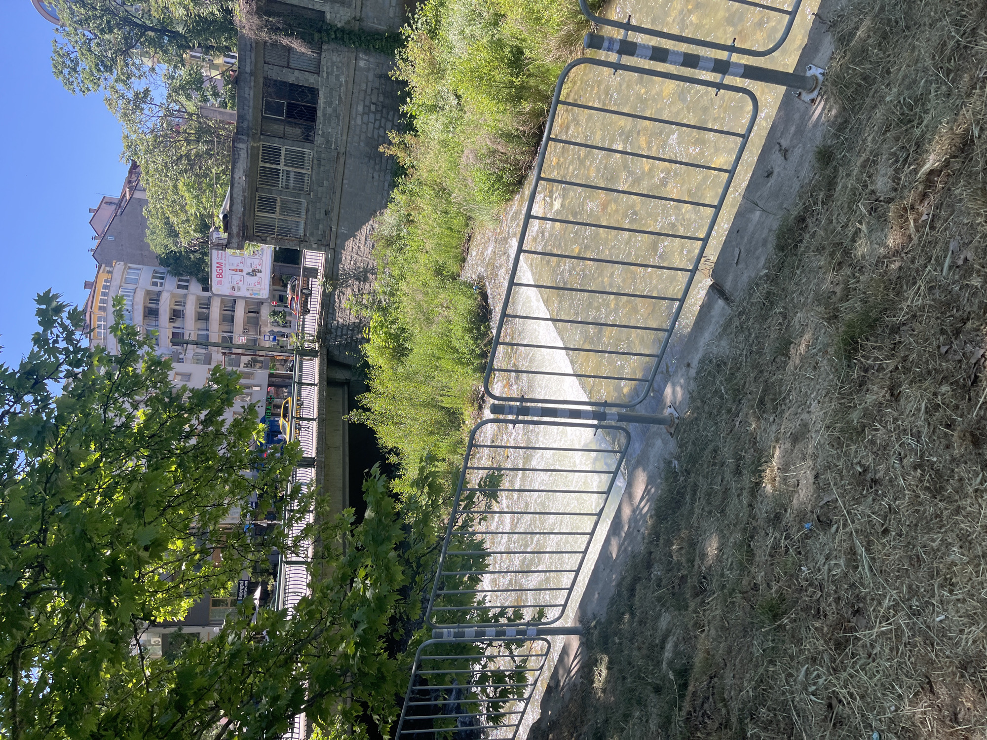 The image shows a metal railing beside a canal in Blagoevgrad, Bulgaria. The canal has flowing water. Vegetation, including trees and grass, flanks the canal. In the background, buildings and a bridge are visible. The sky is clear.