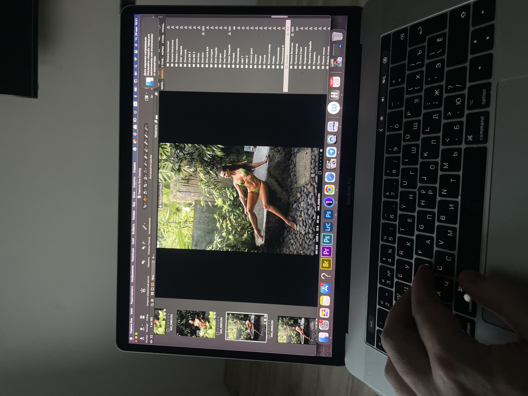 A person is sitting at a desk in a room, working on a Macbook Pro. The laptop displays the Capture One photo editing software with several images of a woman posing in a jungle setting in a bikini. The software window on the laptop is surrounded by a file directory containing several photographs of subjects named Olga and Pavel. The Cyrillic keyboard suggests the location is Russia. The MacBook Pro has several applications pinned to the bottom screen, including Finder, Google Chrome, Safari, Instagram, and Photoshop. The screen also displays the editing software's settings, including ISO, shutter speed, and aperture. A hand is shown typing on the keyboard. The surroundings are dark, with a hint of light coming from the laptop screen. The city of Kazan, Russia, cannot be verified using the data provided.