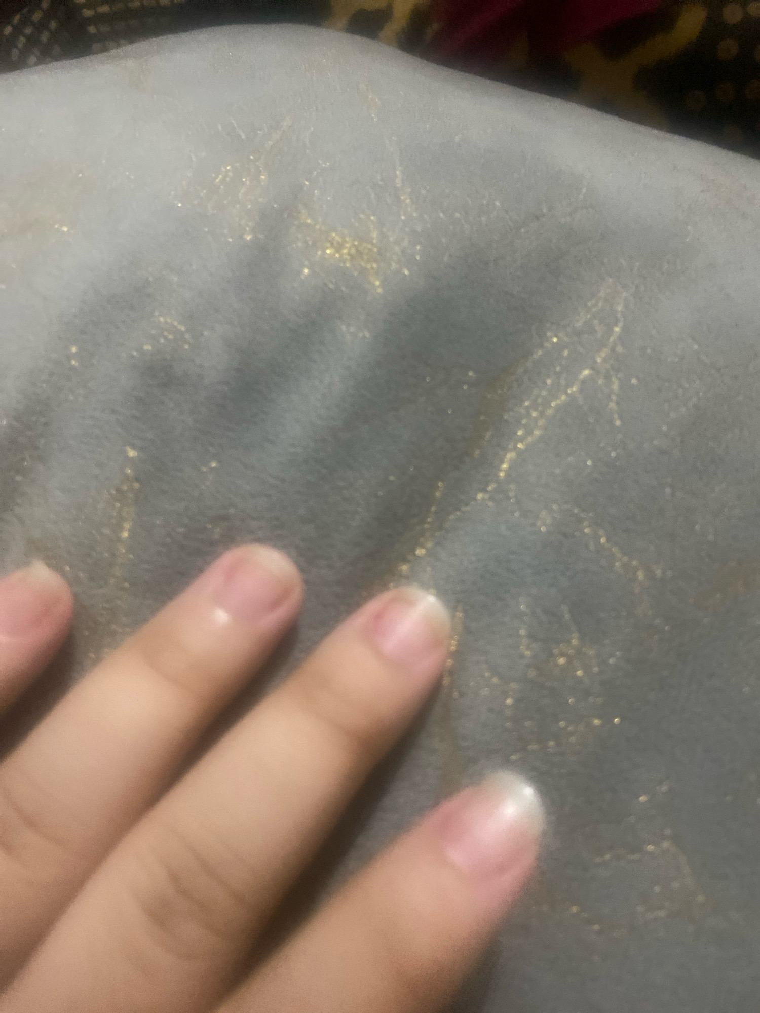 A hand with unpainted fingernails is in the foreground of the image, resting on a textured, gray surface. This surface is flecked with small, shimmering gold particles, some of which form faint, thin lines. The background is blurred and appears to be dark fabric with hints of red and yellow patterns. The scene does not contain enough information to determine its location in Islamabad, Pakistan.