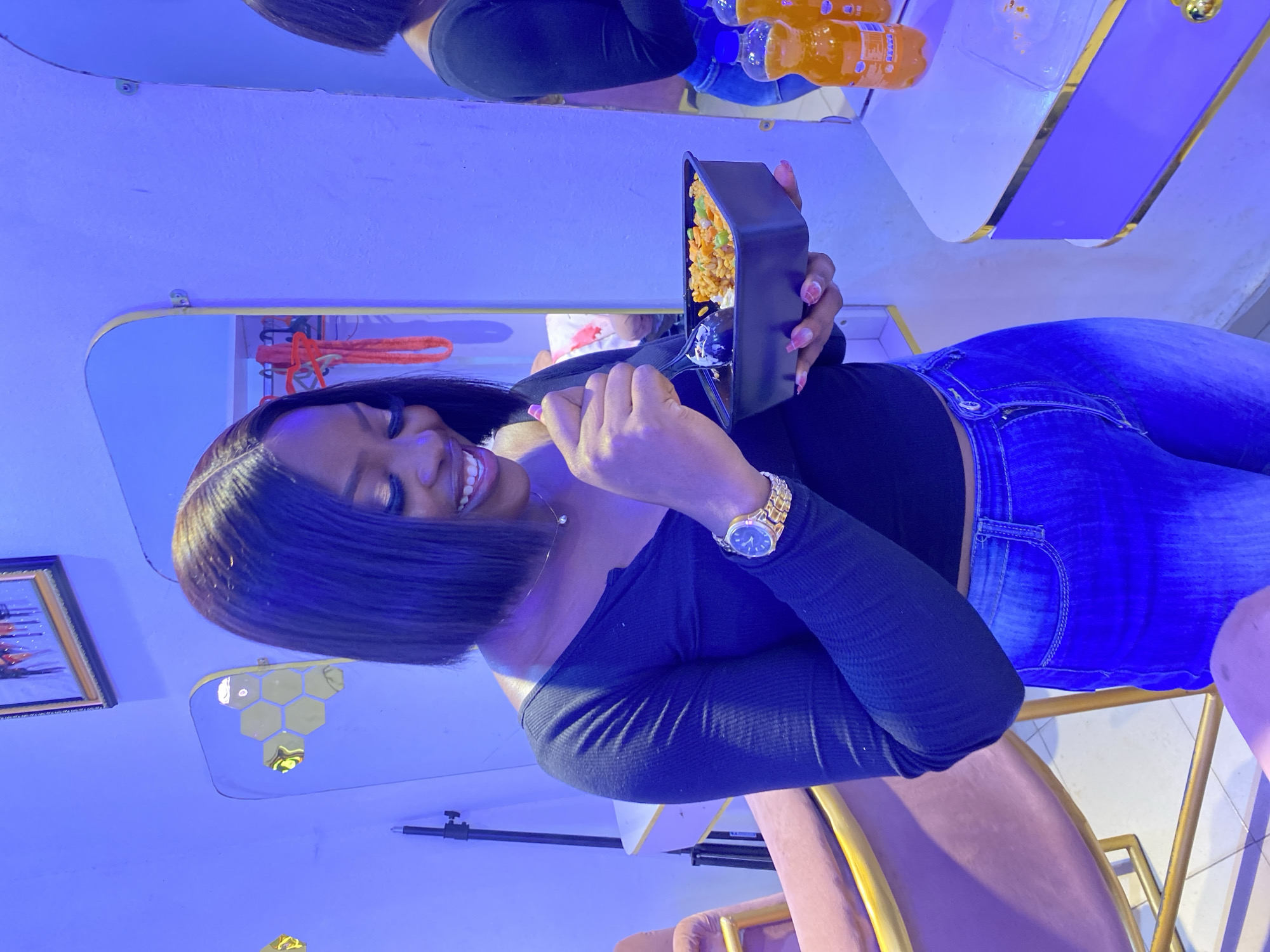 A young woman is depicted in an indoor setting, possibly a cafe or restaurant, characterized by prominent blue-purple mood lighting. She is shown from the waist up, smiling broadly and looking towards the viewer, conveying a joyful and relaxed demeanor. She has dark, straight hair and is wearing a black off-the-shoulder top and blue denim jeans. A gold wristwatch is visible on her left wrist, and her fingernails have light-colored nail polish.
In her right hand, she holds a black rectangular container of food, which appears to be fried rice with visible vegetables like peas and carrots, along with a spoon. Above her, on a shelf or counter, several plastic bottles of an orange-colored beverage are visible. The background features stylish decor, including a mirror reflecting parts of the room, hexagonal decorative elements, and the upper body of another person holding similar beverage bottles. A framed picture hangs on the wall to her left. She is seated in a chair with a purple/pinkish upholstered back and a gold-colored frame. The scene captures a moment of enjoyment, likely during a meal or social gathering, under atmospheric lighting. No discernible text is visible.